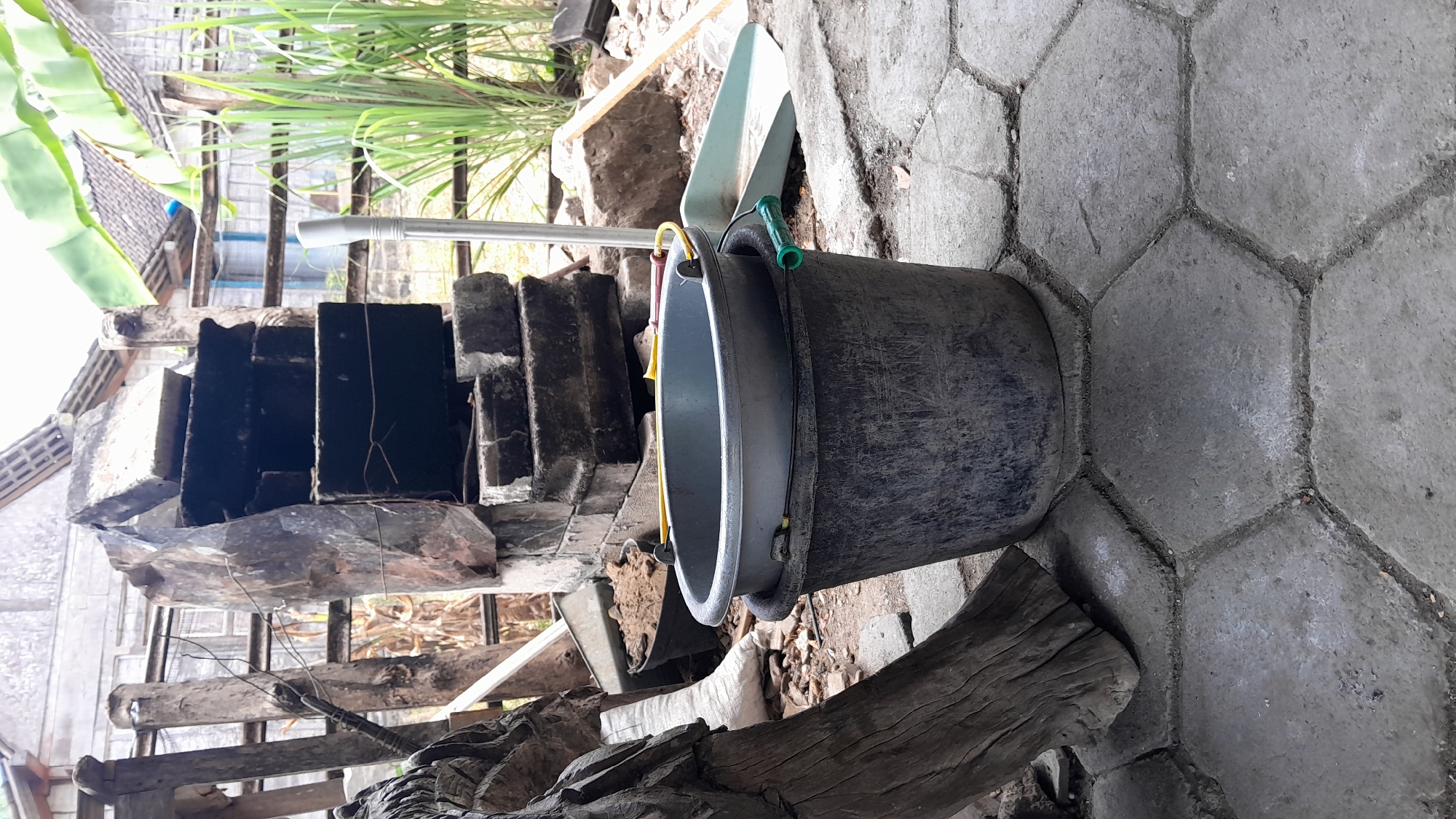 In the city of Sumberarum, Indonesia, a weathered, dark grey plastic bucket sits on a hexagonal concrete paver patio. Inside this bucket, a slightly smaller, lighter grey plastic basin is nested. The basin has a yellow handle. To the right of the buckets, a light green plastic dustpan with a green handle rests on the patio. In the background, a crude outdoor structure made of dark stone and what appear to be concrete blocks is visible. A silver metal pole stands near the stone structure. Beyond that, a wooden fence is present, and lush green vegetation, including what appear to be banana leaves and lemongrass, grows tall. A wooden beam, likely part of a structure or a piece of debris, is positioned to the left of the buckets, leaning against unseen supports. The ground around the buckets is strewn with small stones and dirt. The overall scene suggests an outdoor area, possibly a backyard or workspace, in a rural or semi-rural Indonesian setting.