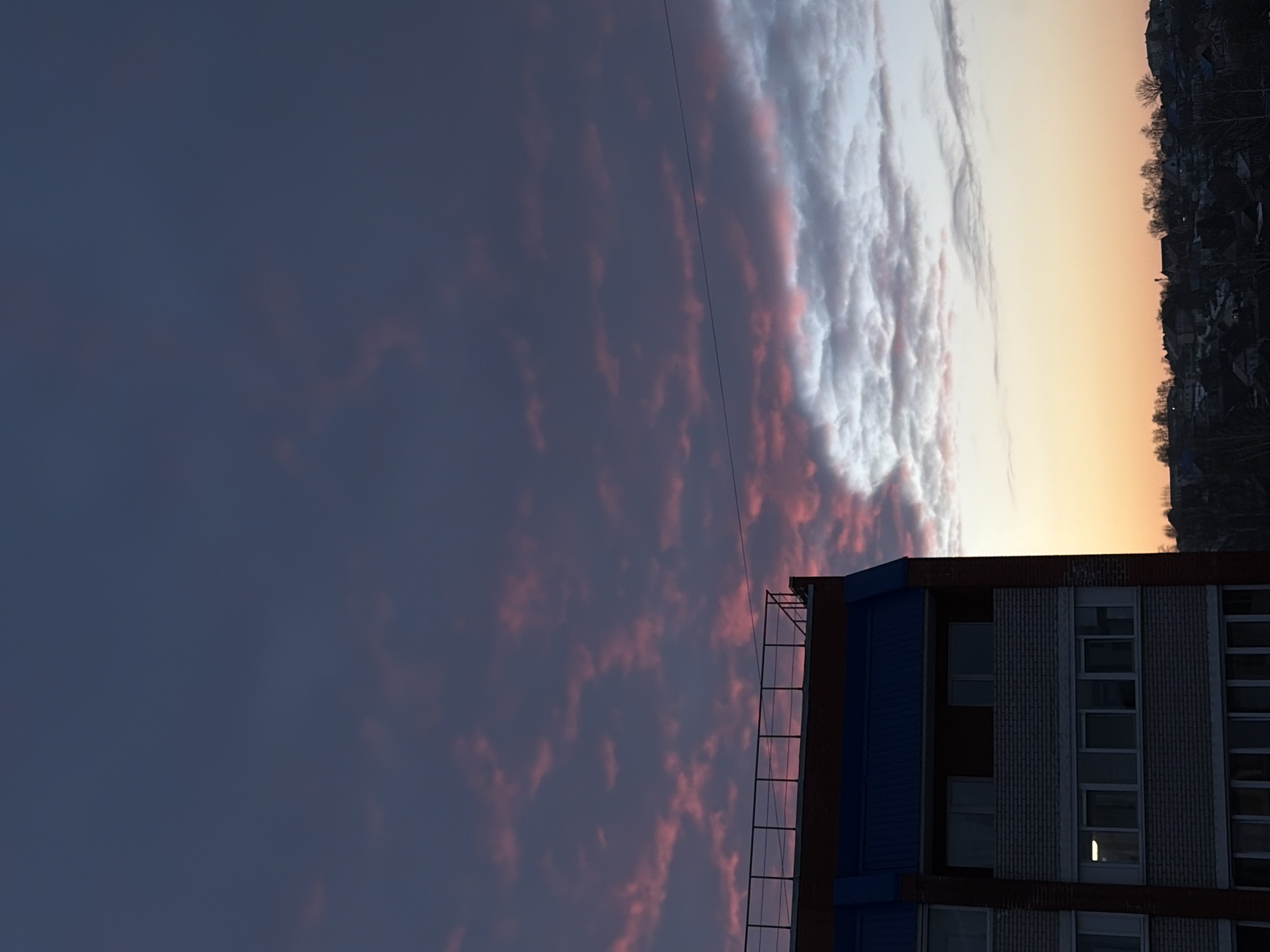 The image depicts an urban-rural transitional landscape at dusk or dawn, centered in Izhevsk, Russia. The sky dominates the upper two-thirds of the frame, exhibiting a deep indigo-purple hue overhead that transitions to pink-tinged clouds across the mid-section, then to an orange-yellow gradient at the horizon on the right. A thin, dark cable or power line traverses horizontally through the mid-right sky.
In the lower left quadrant, a multi-story building occupies the foreground. It features a red-brown brick exterior framing lighter-colored brick sections and rectangular windows. A prominent dark blue sloped roof section is visible below a flat rooftop, which is enclosed by a dark metal railing. One window on the lower visible floor displays an interior light. No individuals or active interactions are observed on or around the building.
The lower right quadrant and horizon reveal a distant residential area composed of numerous houses with dark roofs, interspersed with bare trees. The topography suggests rolling hills. No individuals or active interactions are discernible within this distant landscape.