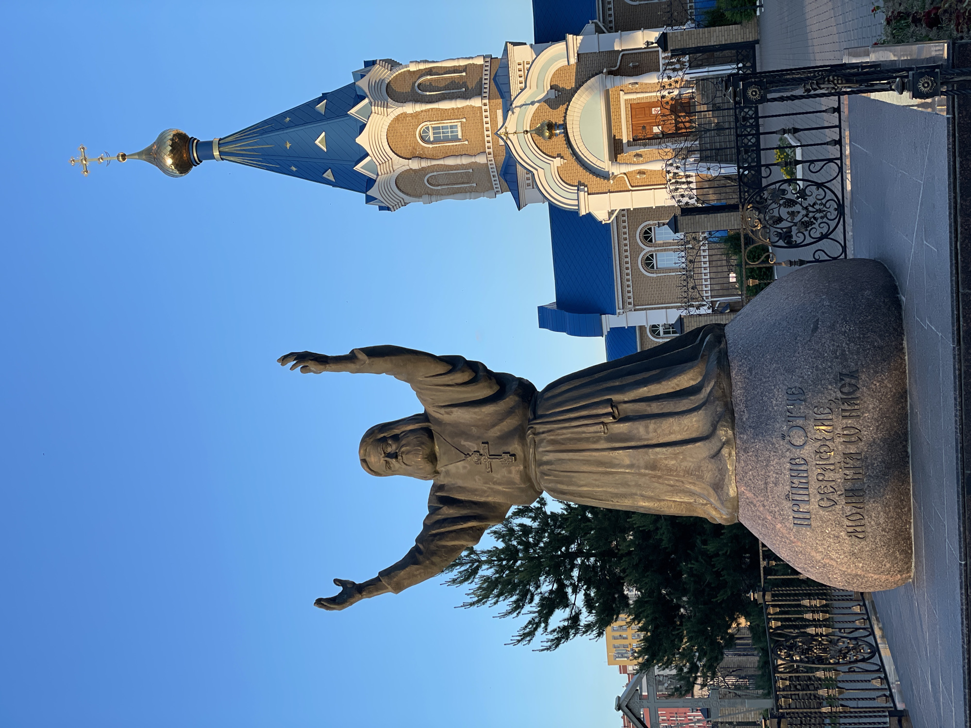 This image, captured in Izhevsk, Russia, depicts a bronze monument of a robed figure, likely a saint, set against a clear blue sky with an Orthodox Christian church in the background.
The central subject is a large, weathered bronze statue of an elderly, bearded man dressed in monastic robes, with a cross pendant visible around his neck. His arms are outstretched in a wide, embracing or blessing gesture, conveying a sense of welcome or benediction. The statue stands on a dark, polished granite base, which features an inscription in Cyrillic script: "ПРЕПОДОБНЫЙ СЕРГИЙ РАДОНЕЖСКИЙ МОЛИТВЕННИК РУСИ." This translates to "REVEREND SERGIUS OF RADONEZH PRAYERFUL INTERCESSOR OF RUSSIA," identifying the figure as Saint Sergius of Radonezh.
Behind the monument, an ornate Orthodox church dominates the middle ground. It features a striking blue-shingled roof with distinctive triangular gables and a prominent golden onion dome topped with a golden Orthodox cross. The church walls are constructed from light-colored brick or stone, with intricate architectural details including arched windows, decorative cornices, and cream-colored accents.
To the right of the statue and extending into the mid-ground is an elaborate black wrought-iron fence or gate, decorated with golden accents and intricate scrollwork patterns, including floral motifs and sunbursts. A lush green tree or large shrub is partially visible behind the statue, adding a natural element to the scene. The foreground shows paved ground, possibly stone tiles. In the far background, hints of other urban buildings are visible.
The clear, vibrant blue sky suggests a bright, sunny day, likely during late morning or early afternoon, with warm light illuminating the church's facade and dome. The overall scene presents a serene and reverent atmosphere, highlighting a significant religious figure and architectural landmark.