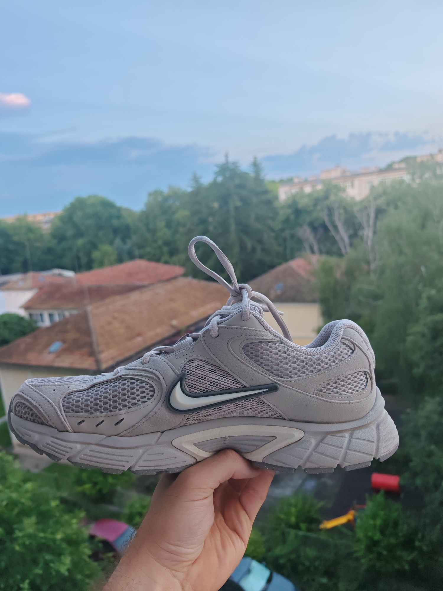 A light grey Nike athletic shoe is centrally positioned, held aloft by a human hand, against a blurred urban background in Shumen, Bulgaria. The shoe exhibits a mesh and synthetic upper in light grey, featuring a white Nike "Swoosh" logo with a black outline on its lateral side. The laces are light grey and untied. The visible hand, positioned beneath the shoe, shows part of a forearm with hair, suggesting a male subject. The deeply out-of-focus background depicts a cityscape with numerous residential buildings characterized by prominent brown and orange tiled roofs, interspersed extensively with green foliage from various trees. The distant background reveals a pale blue sky with indistinct cloud formations. Below the main subject, blurred elements suggest ground-level activity, including partially visible vehicles (one teal, one red) and what appear to be red and yellow components of playground equipment.
