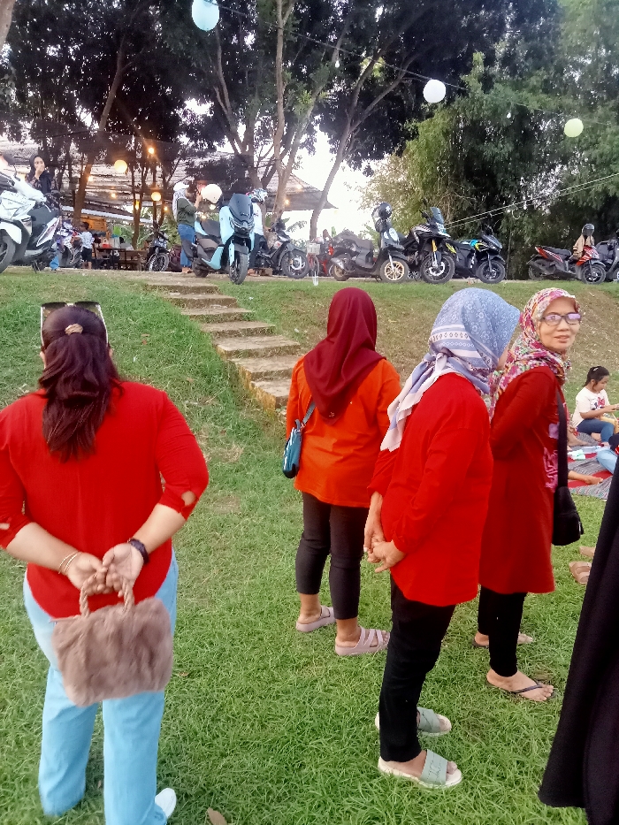 The image shows a group of women standing on a grassy slope outside a cafe in Jakenan, Indonesia (coordinates: -6.758, 111.075). The women are all wearing red clothing and headscarves, except for one woman wearing blue jeans and a red shirt. They are standing in front of a line of parked motorcycles, some of which have riders on them. The scene is bright and sunny, and the trees in the background are green. The women are talking and interacting with each other. The slope is covered in lush green grass, and the environment is calm and relaxed.