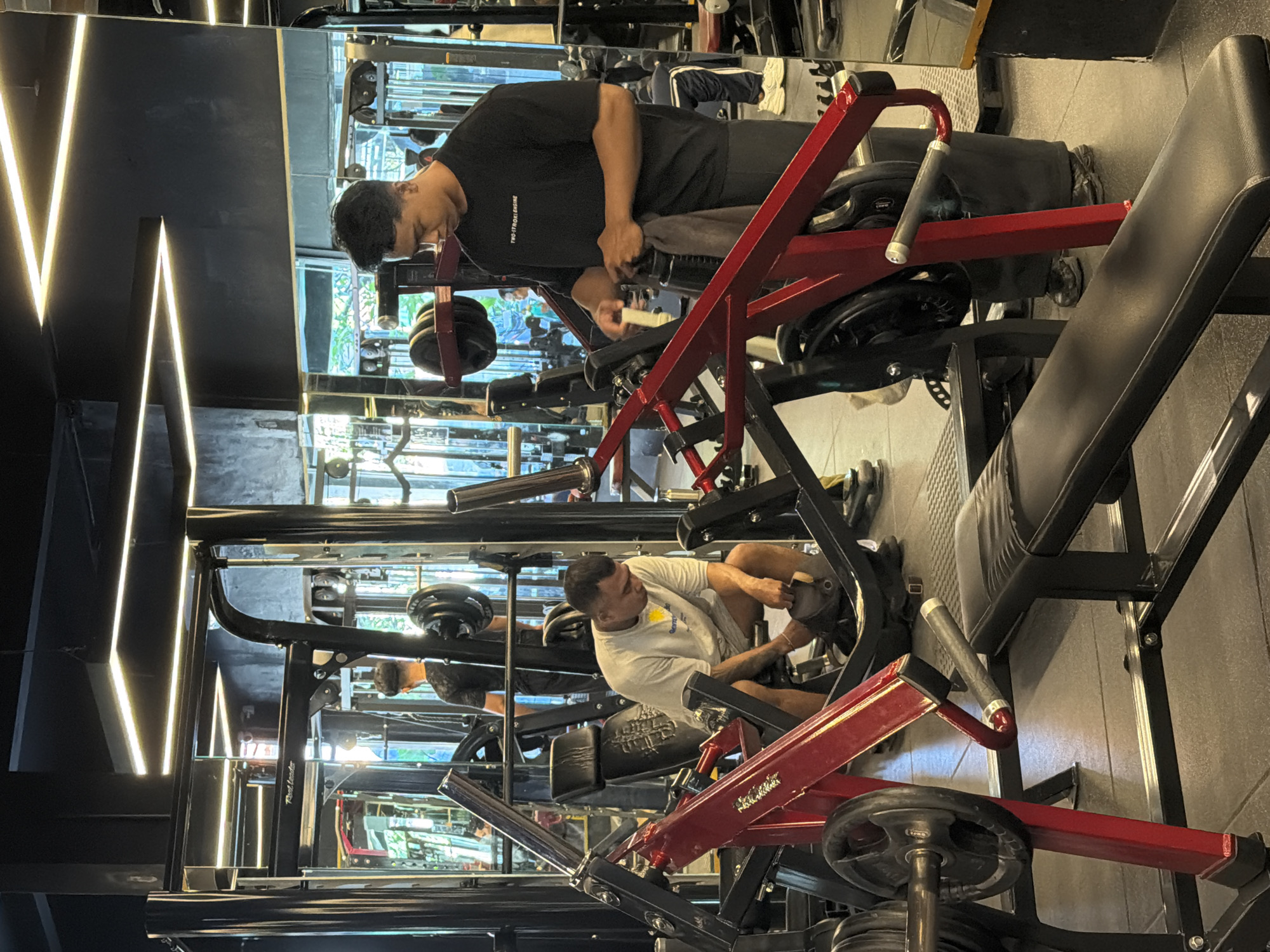 The image shows two men working out on leg press machines in a gym in Denpasar, Indonesia. One man is wearing a black t-shirt and the other is wearing a white t-shirt. Both are adjusting the weights on the machines. The gym has mirrored walls and fluorescent lighting.