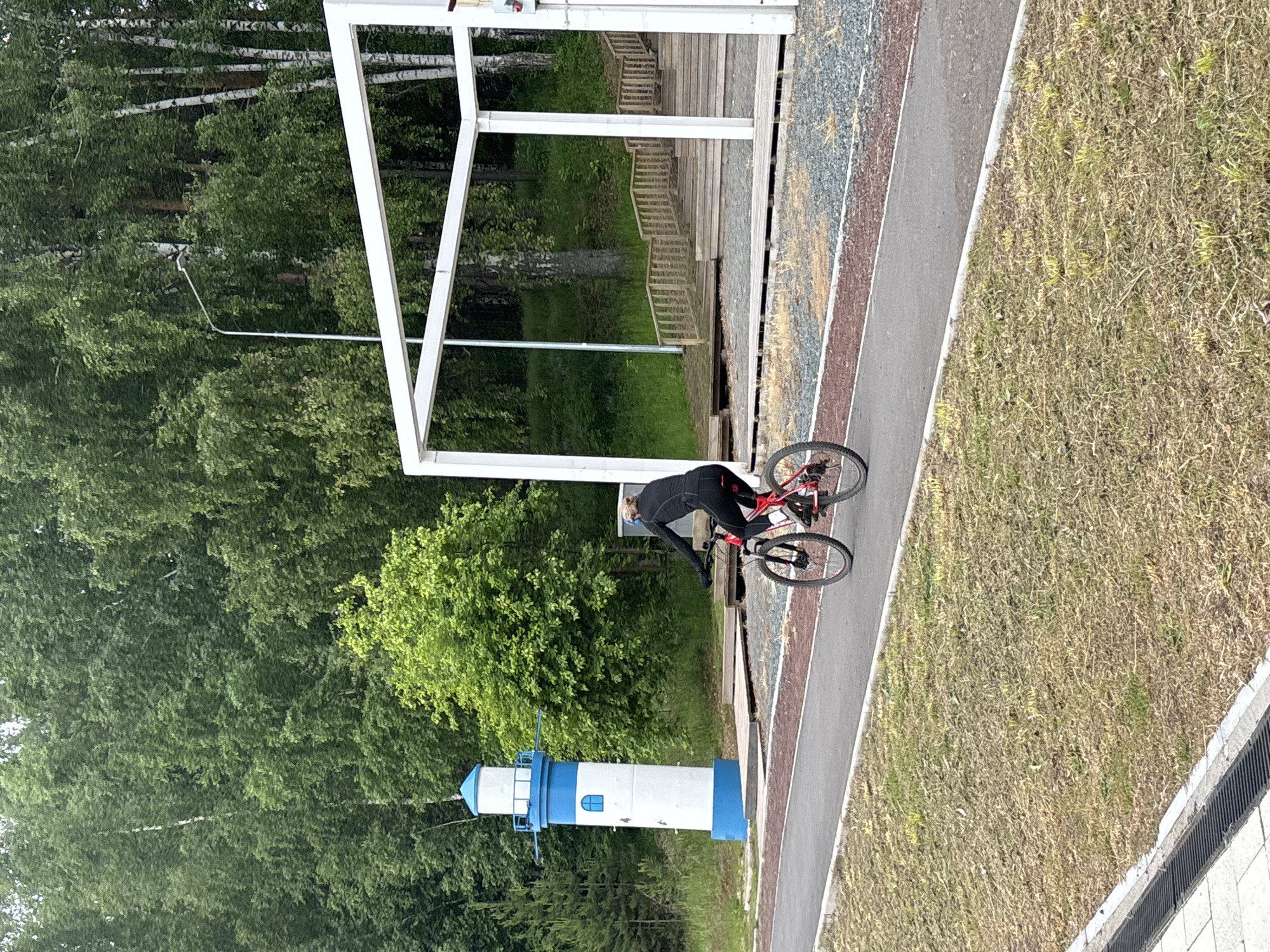 The image depicts an outdoor recreational area in Chaikovsky, Russia, observed during daytime under natural light. A person dressed in dark athletic wear is positioned on a red and black bicycle on a paved asphalt path, suggesting they are either riding or about to ride.
The scene features several elements:
- A clear, grey asphalt path stretches horizontally across the mid-ground, bordered by a reddish-brown strip and a grey gravel area on one side, and dry, yellowish-green grass with a concrete curb and drainage grate on the other.
- To the left of the path, a prominent white architectural frame structure stands, beneath which wooden stairs ascend into a wooded area. This structure might be part of a viewing stand or a park facility.
- In the background and to the left of the path, a striking white and blue lighthouse-like tower is visible amidst dense green foliage. The tower features a blue base, a white cylindrical body with a small blue-framed window, and a blue top section adorned with a spire and horizontal bars.
- The background is dominated by a lush forest of green trees, including several tall birch trees with characteristic white trunks, indicating a natural, possibly extensive, woodland setting.
The overall environment suggests a well-maintained park, cycling track, or a scenic walking trail. The time of day appears to be late morning or afternoon, with soft, diffused light and no harsh shadows. No specific event is clearly defined beyond the presence of the cyclist, and no discernible text or strong emotions are visible.