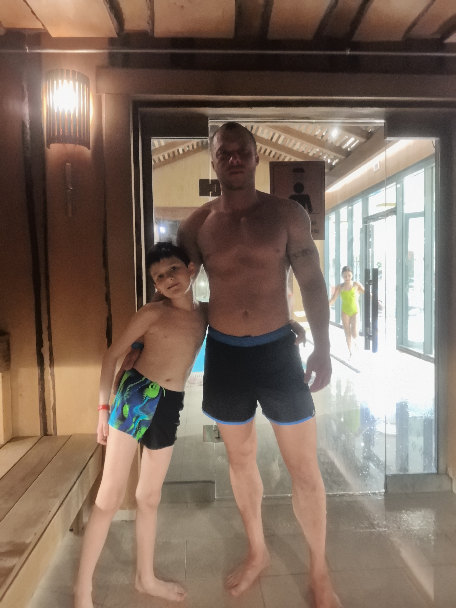 A fair-skinned man with short, dark hair and a shirtless torso poses with a fair-skinned boy of about 8 to 10 years of age in a brightly lit indoor location. The man is wearing dark-colored shorts with a blue trim, and the boy is wearing similar shorts with green and blue accents. The man has his arm around the boy's shoulders. The boy stands slightly in front of the man. Behind them is a glass doorway with a sign indicating a restroom. The doorway leads to a well-lit corridor. A girl in a lime green swimsuit is walking through the corridor. To the left of the man and boy are wooden benches and a wall with a decorative lamp. The floor appears to be wet, suggesting a pool or sauna environment. The location may be in Nizhny Novgorod, Russia.