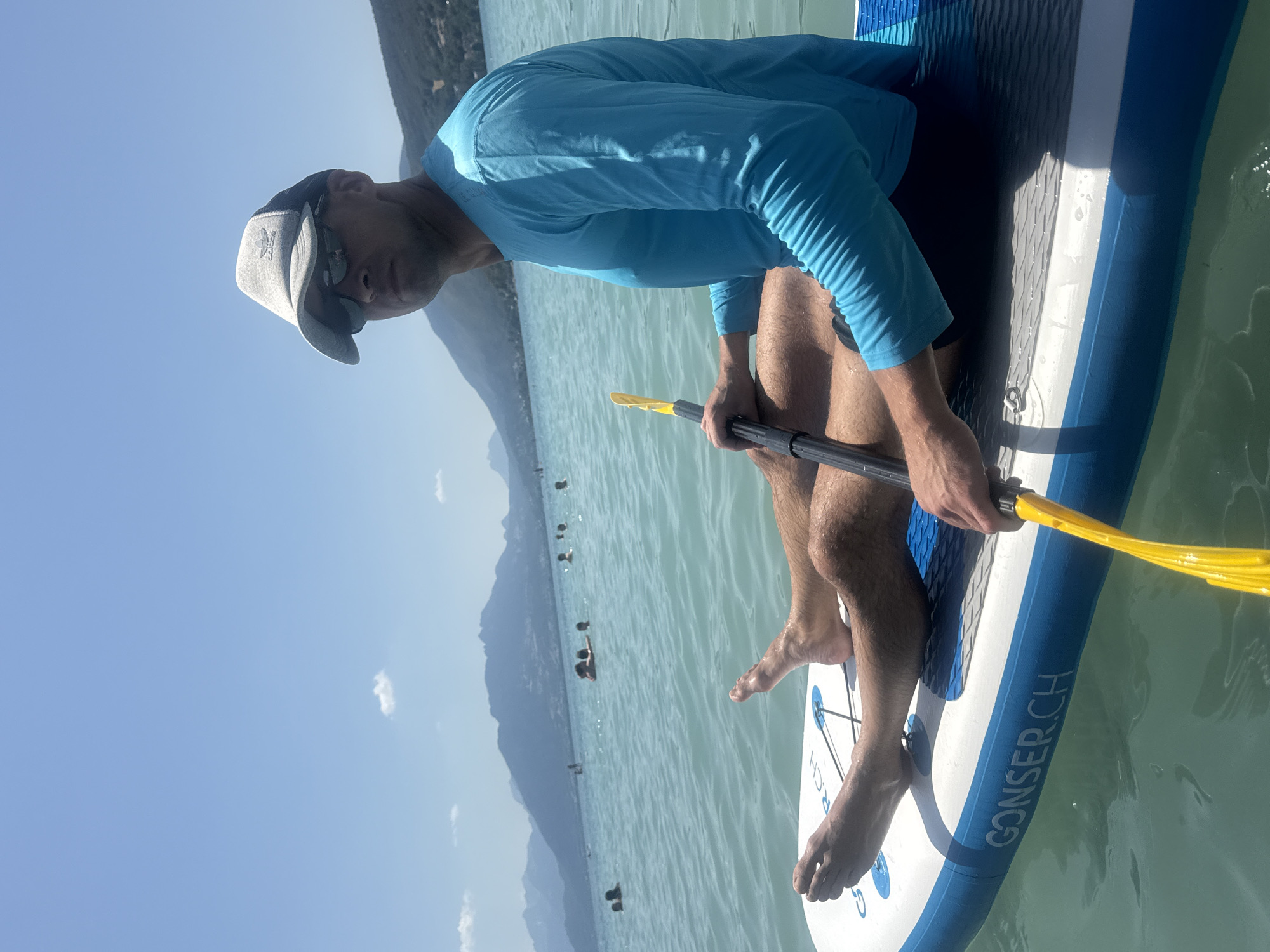 A man is paddleboarding on Lake Annecy, France, under a clear blue sky. He is wearing a blue long-sleeved shirt, black shorts, a grey baseball cap, and sunglasses. He is holding a yellow paddle. The paddleboard is white with blue accents and has the text "CONSER.CH" printed on it. Several other people are visible swimming and enjoying the water in the background, with mountains forming a scenic backdrop. The water is calm and appears to be turquoise.