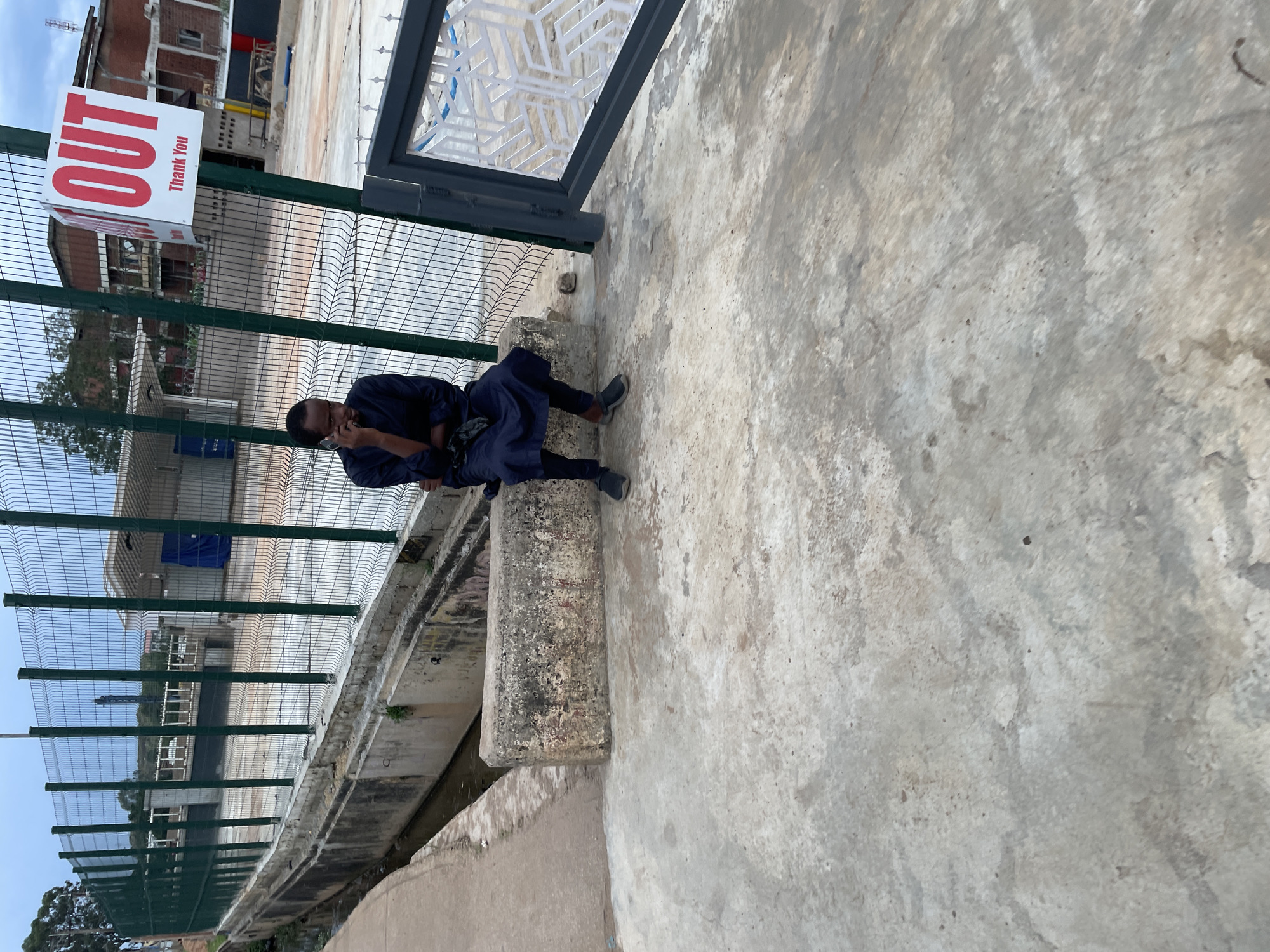The image depicts a street scene in Barakin Lerawi, Nigeria. A Black male, wearing a dark blue outfit and black shoes, is seated on a concrete block next to a drainage channel. He is holding a mobile phone to his ear and appears to be engaged in a conversation. Behind him, a green metal fence encloses an area with buildings and a blue tarp. A red and white sign with "OUT" and "Thank You" is visible attached to the fence. The ground is made of concrete with some visible stains and cracks. The overall environment suggests an urban or semi-urban setting.