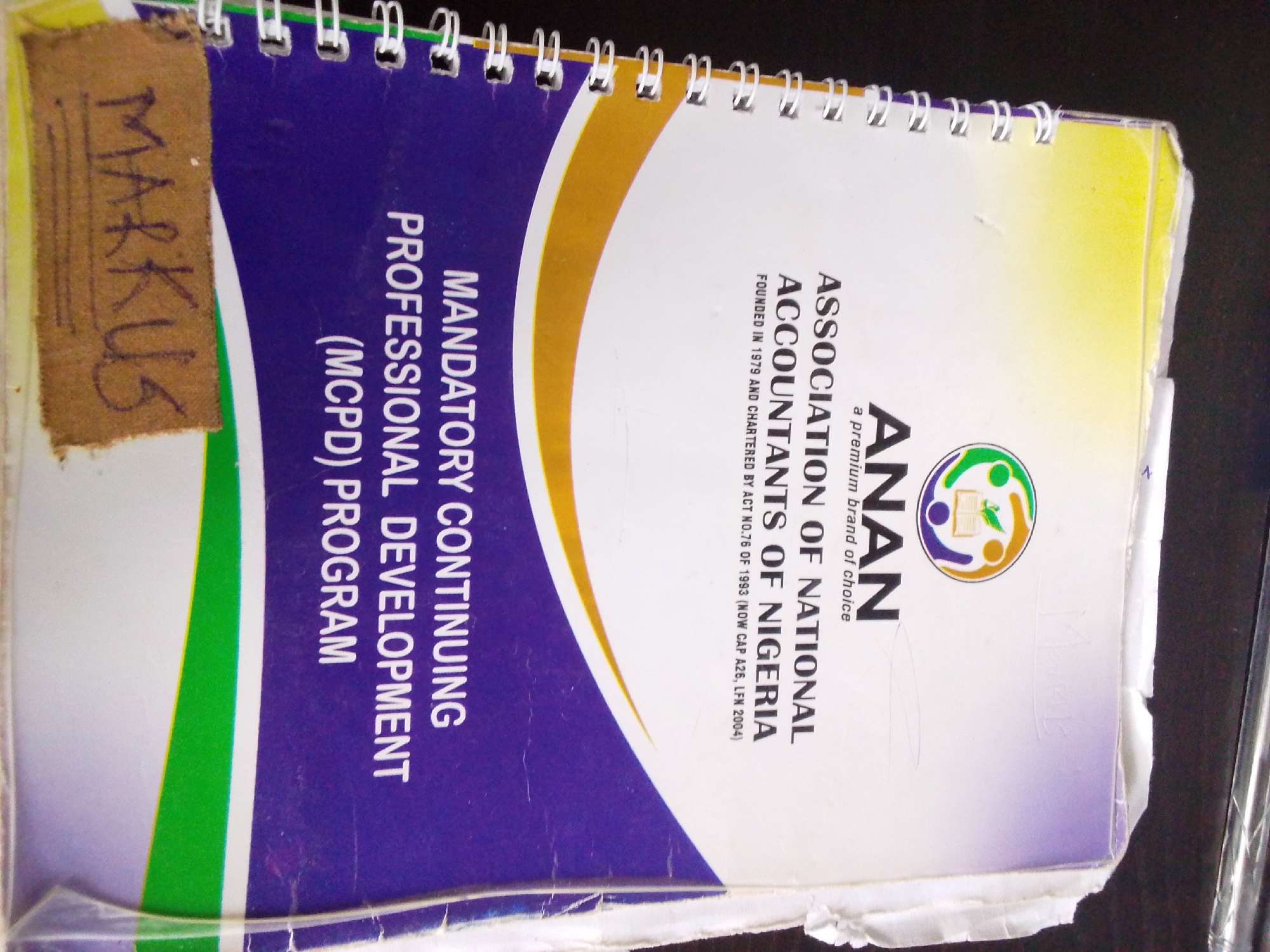 The image is a close-up, low-angle shot of the cover of a spiral-bound book. The book's cover features prominent text that reads "MANDATORY CONTINUING PROFESSIONAL DEVELOPMENT (MCPD) PROGRAM" in large, white, capital letters against a purple background. To the right of this text, there's a graphic design with curved bands of yellow and green. Below the yellow band, the words "ASSOCIATION OF NATIONAL ACCOUNTANTS OF NIGERIA" are printed vertically in white capital letters. Below that, smaller text reads "FOUNDED IN 1979 AND CHARTERED BY ACT NO.76 OF 1993 (NOW CAP A26, LFN 2004)". To the right of this, the logo for ANAN is visible, featuring a stylized circle with figures and a book. On the far left of the cover, a piece of brown paper is taped on, with the word "MARKUS" handwritten in black marker. The image does not provide any information about the city of Kuson, Nigeria, or any people. The environment appears to be indoors, with a dark surface visible in the background.