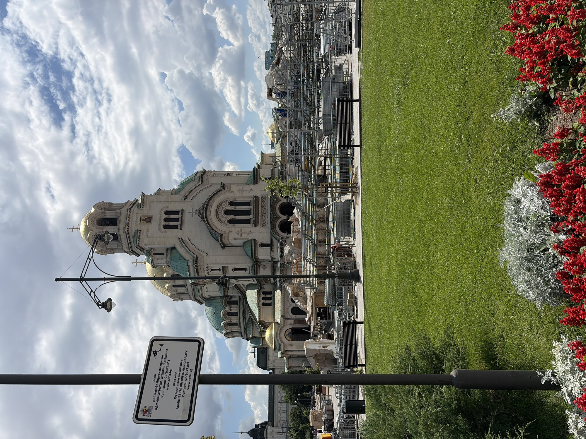 The image depicts the Alexander Nevsky Cathedral in Sofia, Bulgaria, on a partly cloudy day. In the foreground, a well-maintained lawn is bordered by red flowers and silvery-grey foliage. A black lamppost stands to the left of the frame, and a black park bench is situated on a paved walkway behind the lawn.
Dominating the midground and background is the grand Alexander Nevsky Cathedral, characterized by its white facade and golden domes. The cathedral appears to be undergoing renovation or construction, with extensive scaffolding and building materials surrounding its lower sections. Several workers can be seen on the scaffolding.
To the left of the lamppost, a white sign is attached, providing information in both Bulgarian and English. The text indicates "Area with 24 hours video surveillance" and identifies the administrator as "SOFIA MUNICIPALITY," along with contact information and a website.
In the distance, other buildings and structures are visible, including what appears to be another dome on a building to the left of the cathedral. The overall scene captures a blend of historical architecture, urban green space, and ongoing public works.