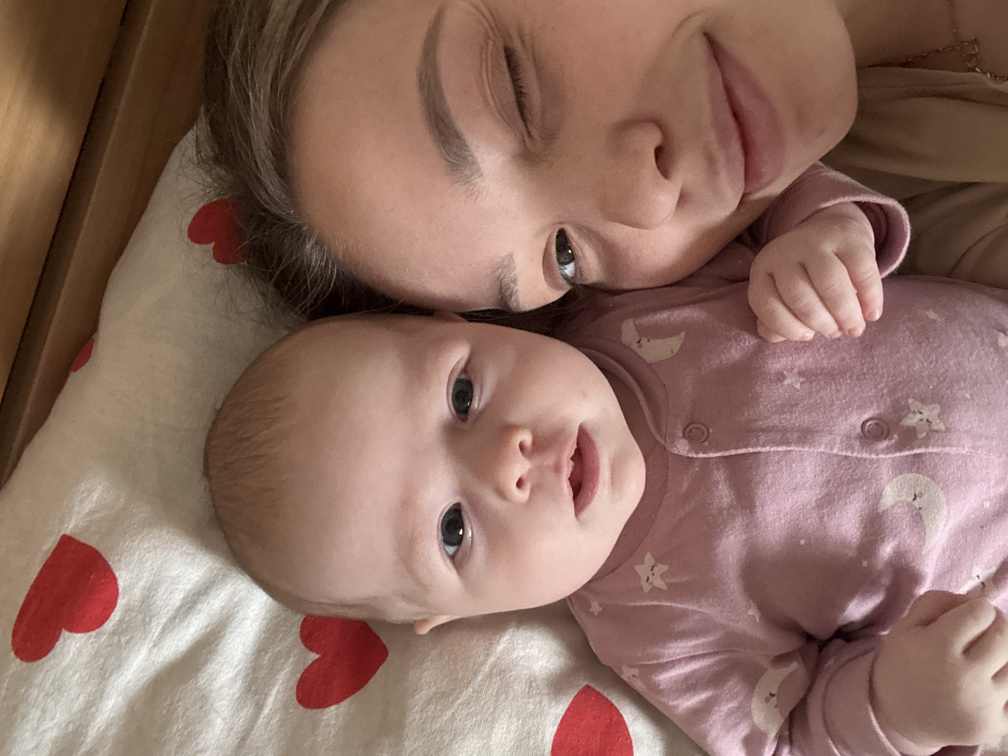 A top-down medium shot features a woman and a baby lying on a bed with a patterned sheet. The woman, fair-skinned, has her eyes partially closed and is smiling slightly. Her head is tilted towards the baby. The baby, also fair-skinned, is wearing a pink onesie with moon and star patterns. They are lying on a white sheet patterned with red hearts. The presence of a wooden structure on the left side of the frame suggests they are in a crib. The photograph was probably taken indoors. The location is unspecified, and there is no evidence to suggest the city of Chaikovsky, Russia.