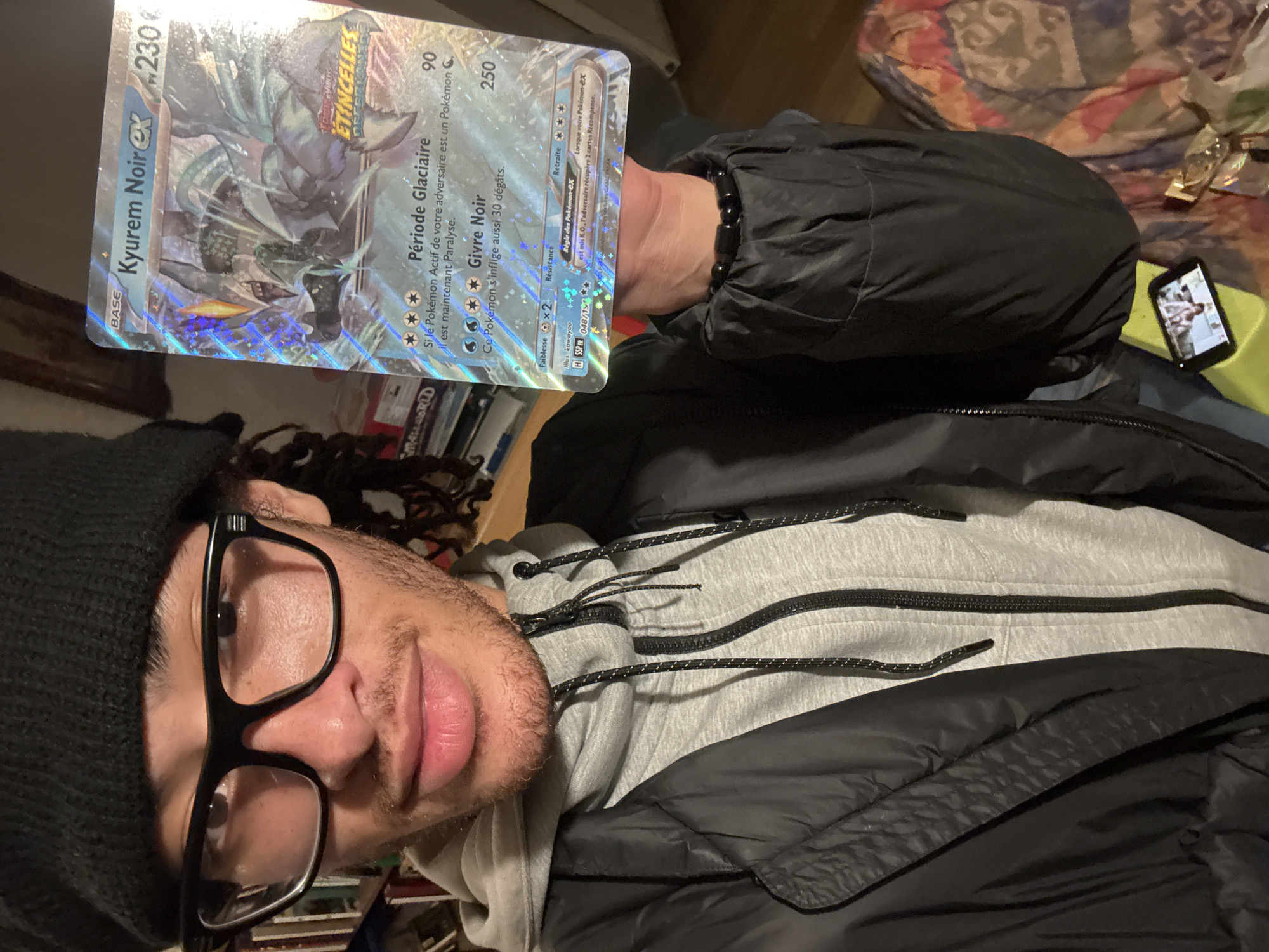 A person with dark curly hair, a black beanie, and black-rimmed glasses is holding a French Pokémon card of “Kyurem Noir EX” in front of them in an indoor setting. The card is silver with the Pokémon’s image and attack descriptions, the card number is visible as "048/115." The person is wearing a grey hooded sweatshirt under a black puffy jacket. A beaded bracelet is visible on their wrist. In the background is a shelf of books and a cell phone.