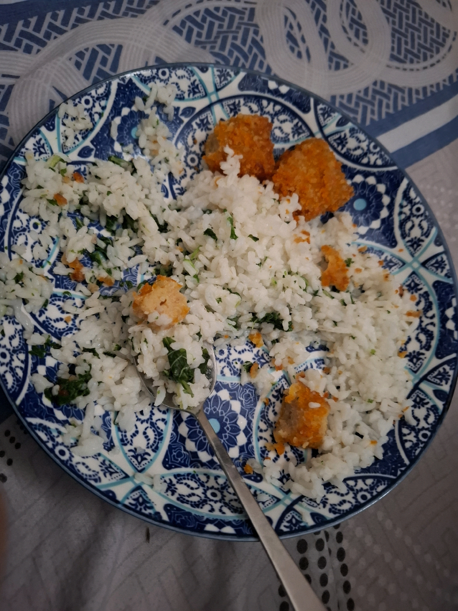 A close-up, overhead shot shows a blue and white patterned plate filled with white rice, green leafy vegetables, and pieces of breaded, fried food. A silver spoon rests on the plate, partially submerged in the rice. The plate is set on a blue and white striped tablecloth. The scene is a still life, likely a meal, with no people visible. The lighting is soft and diffused, suggesting it was taken indoors during the daytime. The image is in focus and exhibits good detail. The location is not discernible from the image itself.