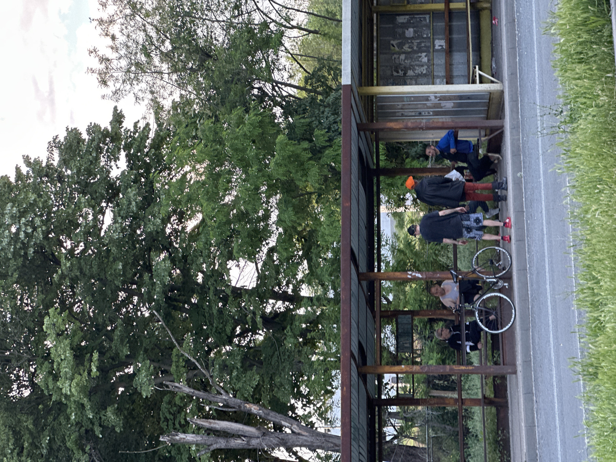 The image, taken outdoors, shows a bus stop or shelter located on the edge of a road. The city is Lovech, Bulgaria. The shelter is constructed of metal and has rusted in places. Some people are inside the shelter. A person wearing a blue shirt reclines on a bench, apparently smoking. Further down the line, a person in an orange hat stands over another seated figure. Another person with a black jacket stands near a bike. A grassy area lines the roadside. Beside the road, trees surround the shelter.