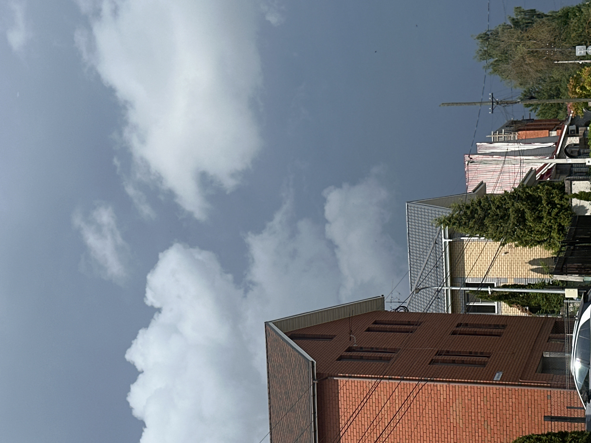 The image captures a residential street scene under a dramatic, overcast sky in Nizhny Novgorod, Russia. In the foreground to the left, a two-story brick house with a dark, multi-faceted roof dominates the frame. Its brickwork is a reddish-brown hue, and several windows are visible. Power lines are strung across its facade. To the right of this house, a narrower, lighter-colored house with a gray tiled roof is partially visible. Further down the street, other houses with varied architectural styles and roof colors are seen, including one with a pink and white striped facade and another with visible brickwork. Evergreen trees and other foliage are interspersed between the buildings, casting shadows on the ground. A black car is parked in the lower left corner. Utility poles with electrical wires are also present. The sky is filled with a mix of dark gray storm clouds and brighter white, fluffy clouds, suggesting an impending or passing weather event. The overall impression is one of a suburban or semi-urban residential area on a cloudy day.