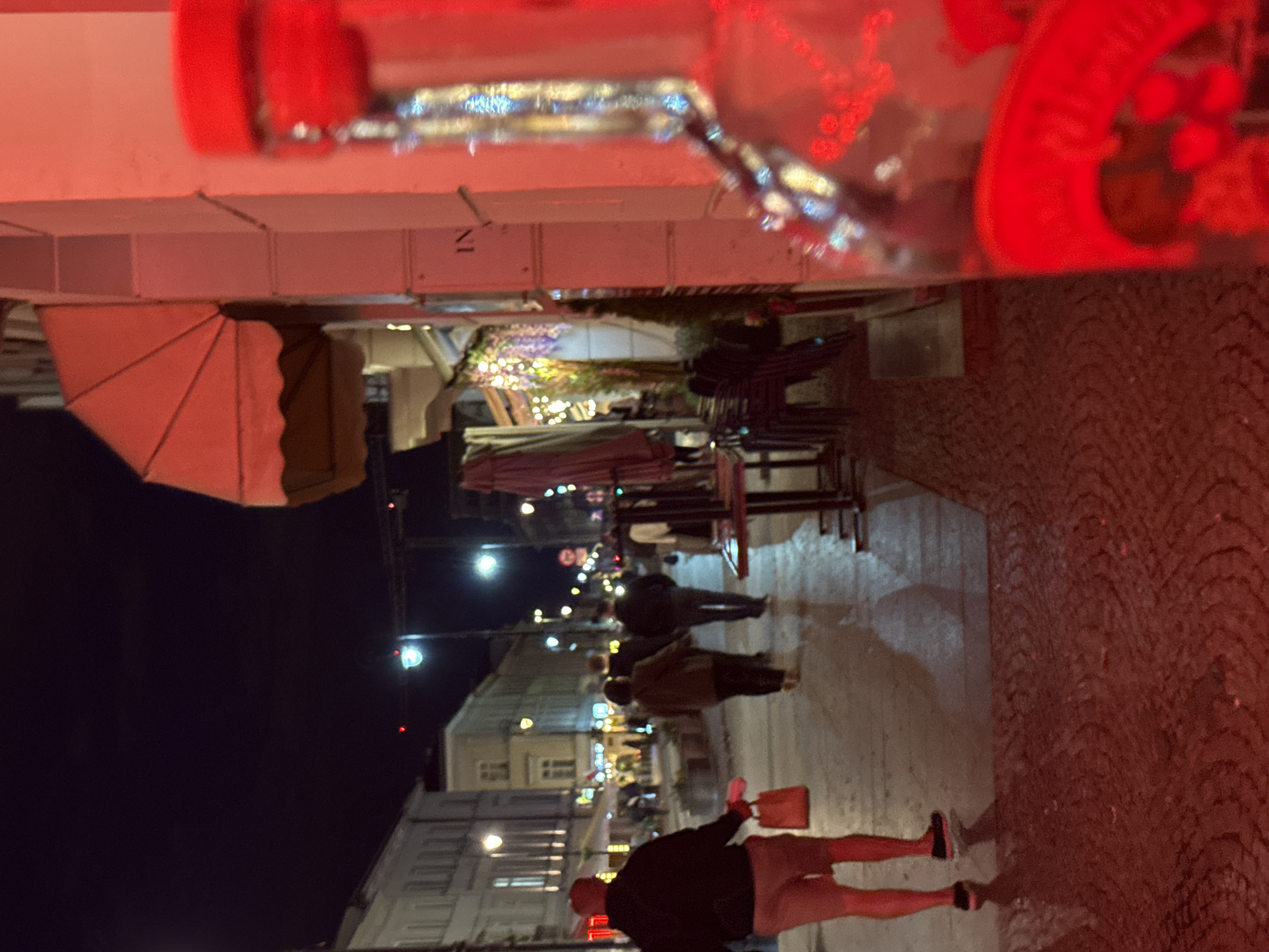 An outdoor scene at night shows a pedestrian walkway with people walking, and a storefront with outdoor seating. In the foreground, a glass bottle with a red cap is blurred and appears to be leaning against a light-colored wall. The bottle reflects colorful lights. A sign on the wall reads "IN." To the left, a person wearing a dark hoodie and red shorts is walking away from the camera, carrying a red shopping bag. Several other individuals are walking further down the street, which is lined with buildings and illuminated by streetlights and decorative lights. There are also tables and chairs for outdoor dining. The ground is paved with cobblestones.