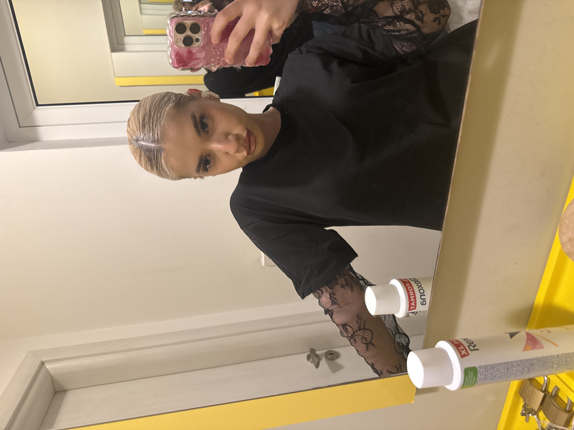 A young woman with blonde, slicked-back hair is captured taking a mirror selfie indoors. She is visible from the chest up, holding a smartphone with a pink, floral-patterned case featuring multiple camera lenses. She wears a black t-shirt layered over a black lace long-sleeve top, with the intricate lace pattern visible on her arms. Her expression is neutral as she looks directly at her phone. Her hair appears wet or recently styled, pulled back from her face, and her darker roots are noticeable.
The mirror in which she is reflected has a prominent bright yellow frame. To the right of the woman's reflection, on a matching yellow surface or shelf, are two white bottles that resemble hair care products and a small collection of silver-colored padlocks. One bottle clearly shows "Rex" and "XL", while the other has partially mirrored Cyrillic text, including what appears to be "ТАМБОУ" (Tambov) and other words likely related to hair products or color, such as "блэндая" (blonde).
The reflection also reveals a white window frame to the left and a light-colored wall or ceiling in the background. The setting suggests a personal space, possibly a bathroom or a room used for hair styling. The location is Sofia, Bulgaria.