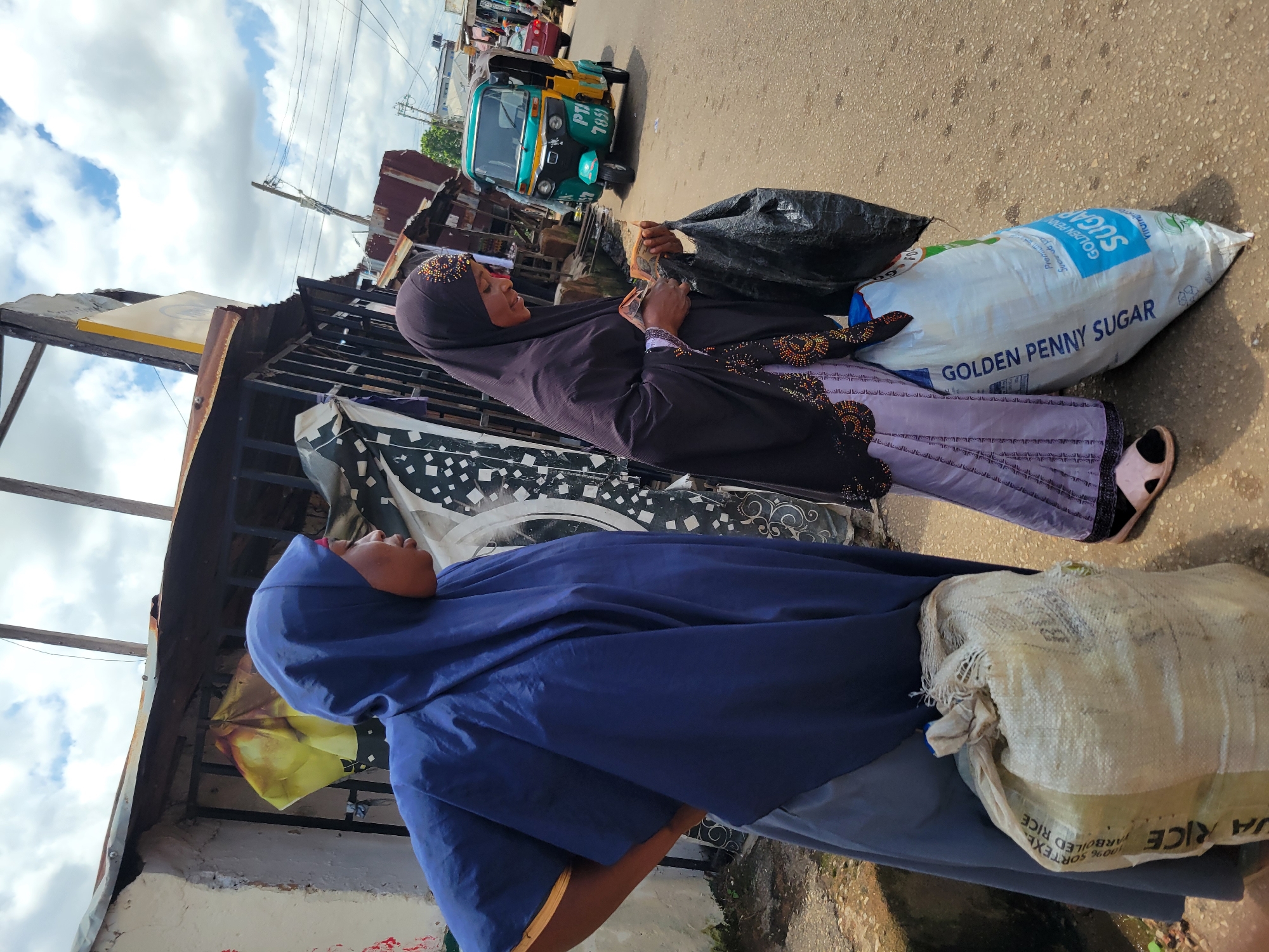 In Jos, Nigeria, two women are pictured in what appears to be a market or roadside setting. The woman on the left wears a dark blue hijab and a light blue abaya, with a large, off-white woven sack at her feet. The woman on the right wears a dark brown hijab and a patterned lavender dress. She is holding a black plastic bag in her left hand and counting money in her right hand. In front of her is a large, white sack with "GOLDEN PENNY SUGAR" printed on it in blue. Behind the women, market stalls with corrugated iron roofs are visible, along with a turquoise and yellow auto-rickshaw. The ground is paved with asphalt. The sky is partially cloudy.