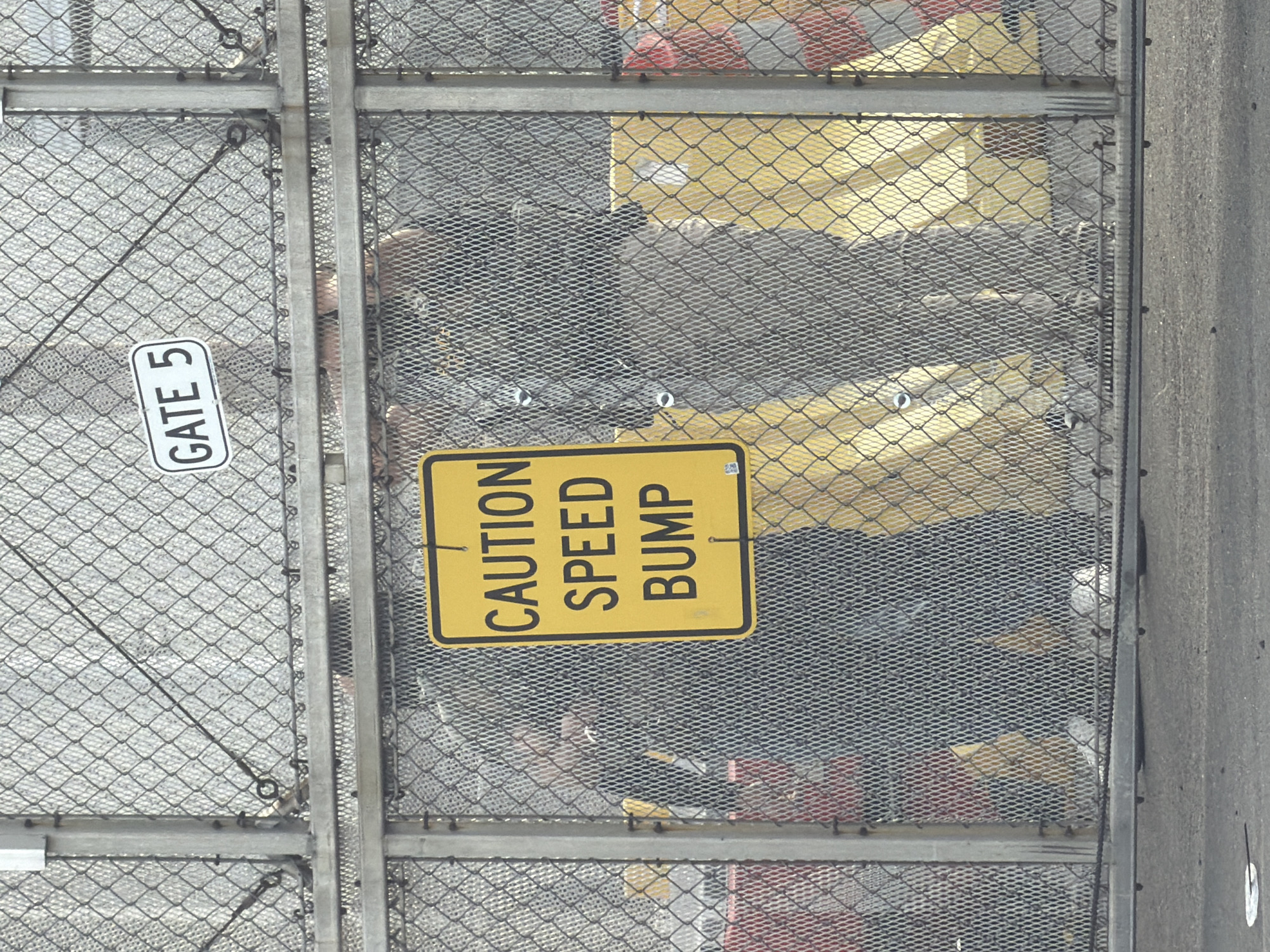 The image shows a chain-link fence with a "GATE 5" sign attached to the left side. A "CAUTION SPEED BUMP" sign is affixed to the fence's right side. Behind the fence, there are indistinct shapes, including hints of yellow, gray, red, and black, possibly representing objects or clothing. The location is the city of Newark, United States of America.