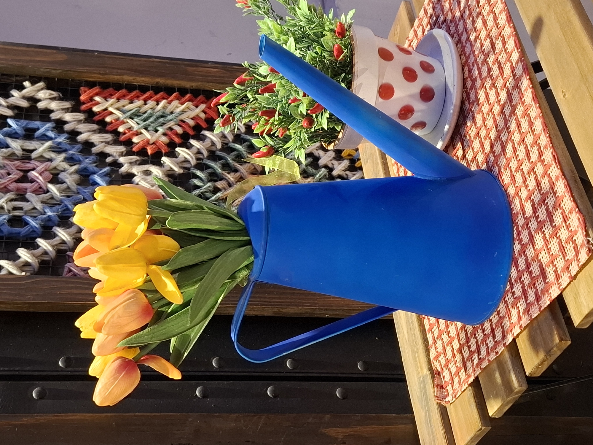 A still life photograph shows a composition of objects arranged on an outdoor surface, likely in Novi Sad, Serbia, given the location request. The objects are placed on a wooden surface covered partially by a red and white checkered cloth. A blue watering can, filled with yellow and orange tulips, is the central element. Beside it, a white pot with red polka dots holds a small artificial plant bearing tiny red peppers. In the background, there's a framed woven artwork featuring patterns in red, white, blue, and green threads. The lighting suggests it is daytime.