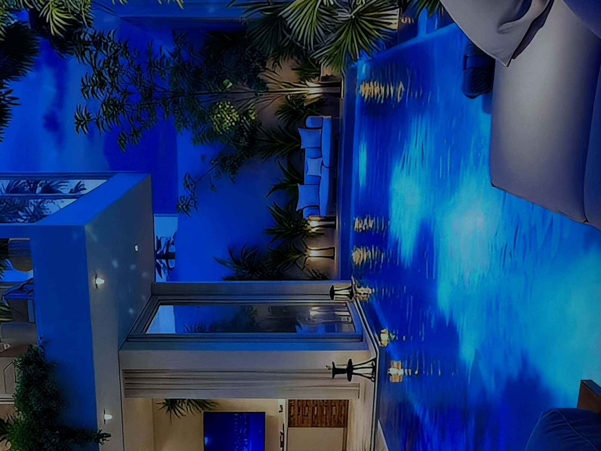 The image captures a luxurious, modern architectural space illuminated by a dominant, vivid blue light at night. The scene features a multi-level building, a large swimming pool or water feature, and lush vegetation, creating an atmosphere of sophisticated tranquility.
The contemporary building showcases clean lines, light-colored walls, and expansive glass windows. Through these windows, glimpses of the interior reveal a television screen glowing blue and wooden slatted wall decor. Several warm-toned accent lights are integrated into the exterior walls, providing a subtle contrast to the prevailing cool blue.
A striking, intensely blue illuminated swimming pool or water feature occupies a significant portion of the foreground and midground. The water's surface displays shimmering reflections of the surrounding architecture and lights, creating dynamic patterns and enhancing the ethereal blue glow. Beside the pool, light-colored outdoor furniture, including a sofa with cushions and another padded lounge piece, is visible, suggesting a comfortable area for relaxation.
The area is richly landscaped with a variety of tropical or subtropical plants, such as palm trees and dense bushes, which are strategically placed around the building and pool, contributing to an exotic and private ambiance.
The setting is distinctly nighttime, emphasized by the strong artificial illumination and the dark background. No people are visible, lending a serene and uninhabited quality to the luxurious environment. The overall mood is calm, inviting, and opulent. There is no visible text in the image.