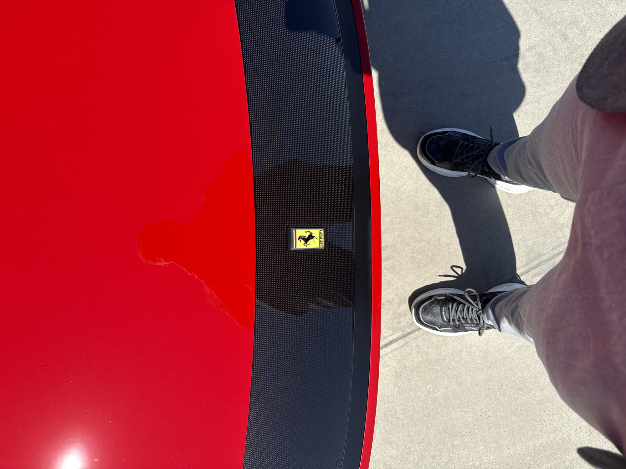 A top-down shot shows a portion of a red Ferrari with a carbon fiber accent down the middle, featuring the Ferrari logo. To the right are a person's legs wearing grey sweatpants and black sneakers. The scene is set on concrete, presumably a street or sidewalk. It is not possible to ascertain the location as Plovdiv, Bulgaria, or any other specific location.