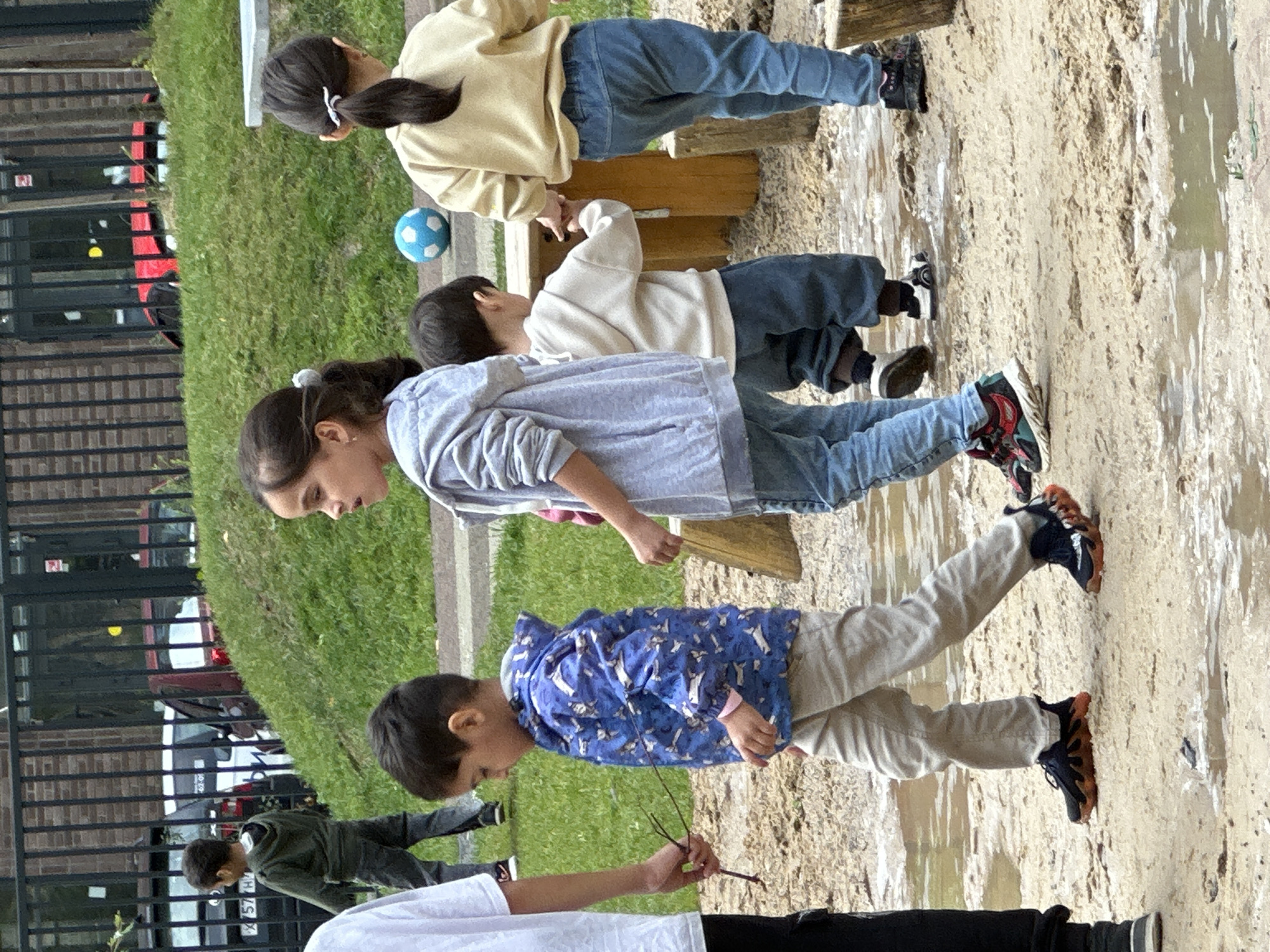 In an outdoor sandpit area, likely at a playground or school in Znamya Oktyabrya, Russia, several children are interacting. A young boy in the foreground wears a blue jacket with a white pattern and tan pants, holding a small stick. To his right, a girl in a grey hoodie and blue jeans walks, looking to her left. Behind her, another child in a cream-colored hoodie and blue jeans is visible, holding hands with a fourth child who is partially obscured by the girl in grey. Further in the background, a young boy in a green hoodie and dark pants walks on a grassy slope. A blue and white soccer ball rests on the ground near a wooden structure. The environment includes a metal fence in the background, with cars and a building visible beyond it, suggesting an urban or suburban setting. The ground is sandy with several puddles of water, indicating recent rain or a water feature in the play area.