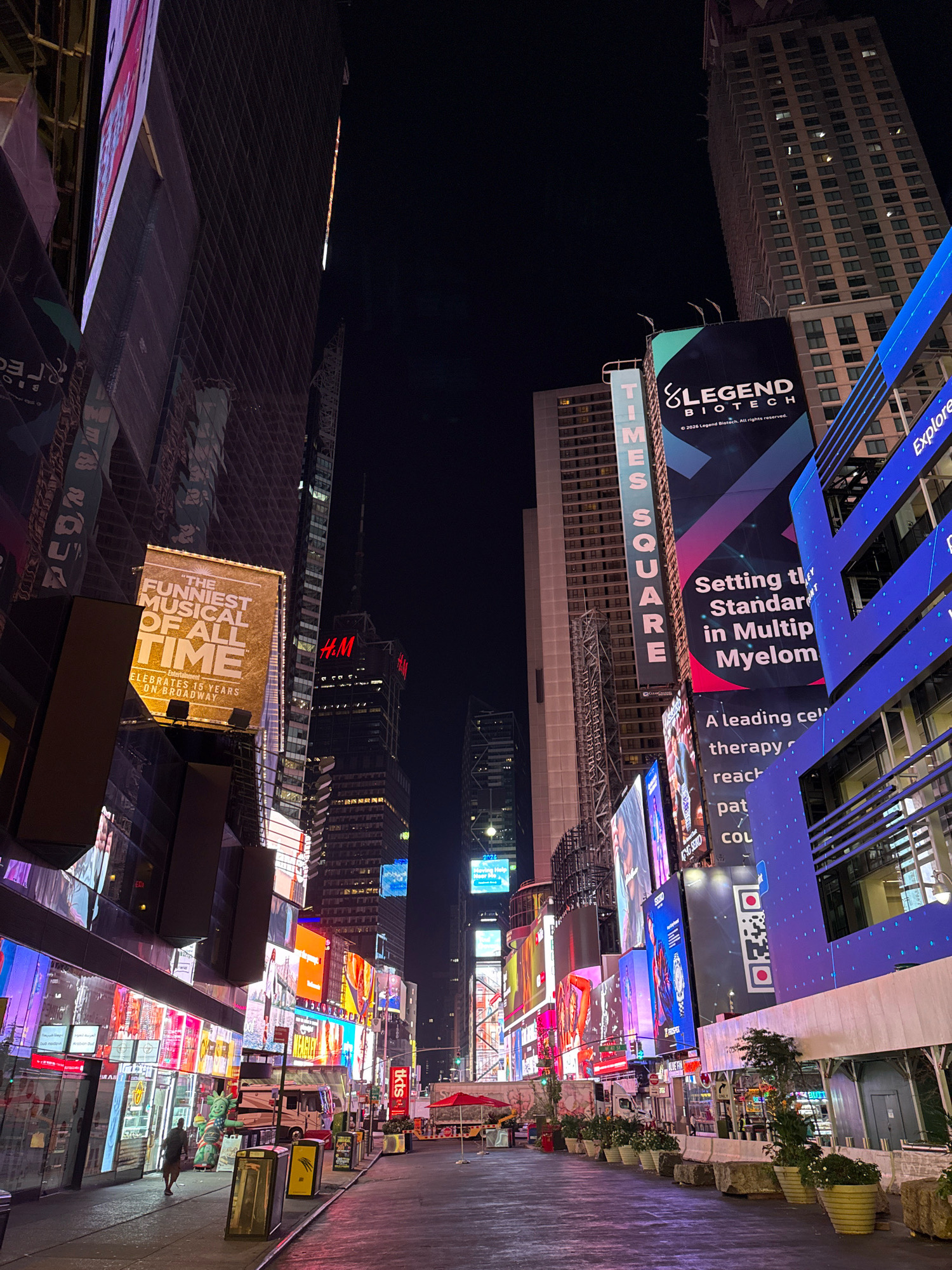 This is a nighttime vertical shot of Times Square in New York City, USA. Towering buildings with numerous illuminated billboards dominate the scene. A large billboard for "Legend Biotech" is prominent, displaying text about setting the standard in Multiple Myeloma. Other billboards advertise "The Funniest Musical of All Time" and "H&M". The street is mostly empty, with a few pedestrians visible. A food cart, some portable barriers, and planters with greenery line the sidewalk. The ground reflects the vibrant lights of the advertisements, creating a colorful and dazzling atmosphere.