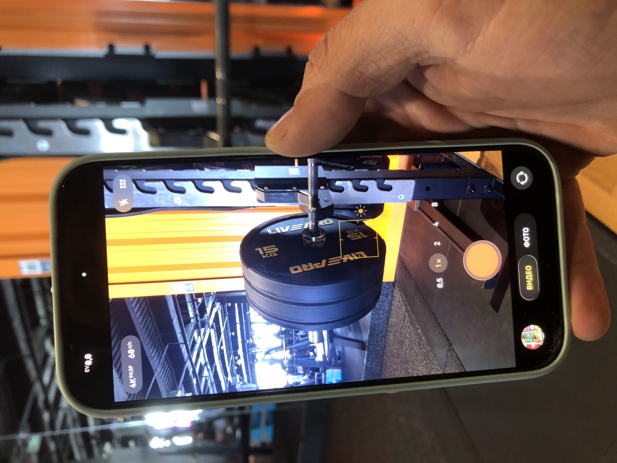 A person's right hand holds a light green-cased smartphone, likely an iPhone given the interface, in a vertical position. The phone's camera application is active, displaying a live preview of an indoor gym setting. The primary subject captured on the phone's screen is a black circular weight plate, prominently displaying "15 KG" and the brand "LIVE PRO" in gold lettering. This weight plate is attached to a piece of black gym equipment, which appears to be a barbell or weight tree. The background visible through the phone's camera shows more gym apparatus, including structural elements and orange beams, illuminated by indoor lighting.
The phone's camera interface is visible on the screen. On the left side, "EV0,0" and "4К РАЗР 60 к/с" (4K RESOLUTION 60 fps) are displayed, indicating the current video recording settings. Various small icons, including a grid and what appears to be a flash setting, are also present. On the right side of the screen, mode selectors for "ВИДЕО" (VIDEO) and "ФОТО" (PHOTO) are shown, with "ВИДЕО" highlighted, confirming the camera is in video mode. Zoom options "0,5", "1x", and "2" are positioned above a large, circular orange shutter button.
The actual background behind the smartphone in the primary image also shows elements of a gym, consistent with the scene being captured by the phone. The person's thumb is visible near the top edge of the phone. The overall setting is an indoor gym, and based on the Cyrillic text on the phone screen, the location is Krasnoyarsk, Russia. The image captures the moment of a user interacting with their phone's camera in a fitness environment.