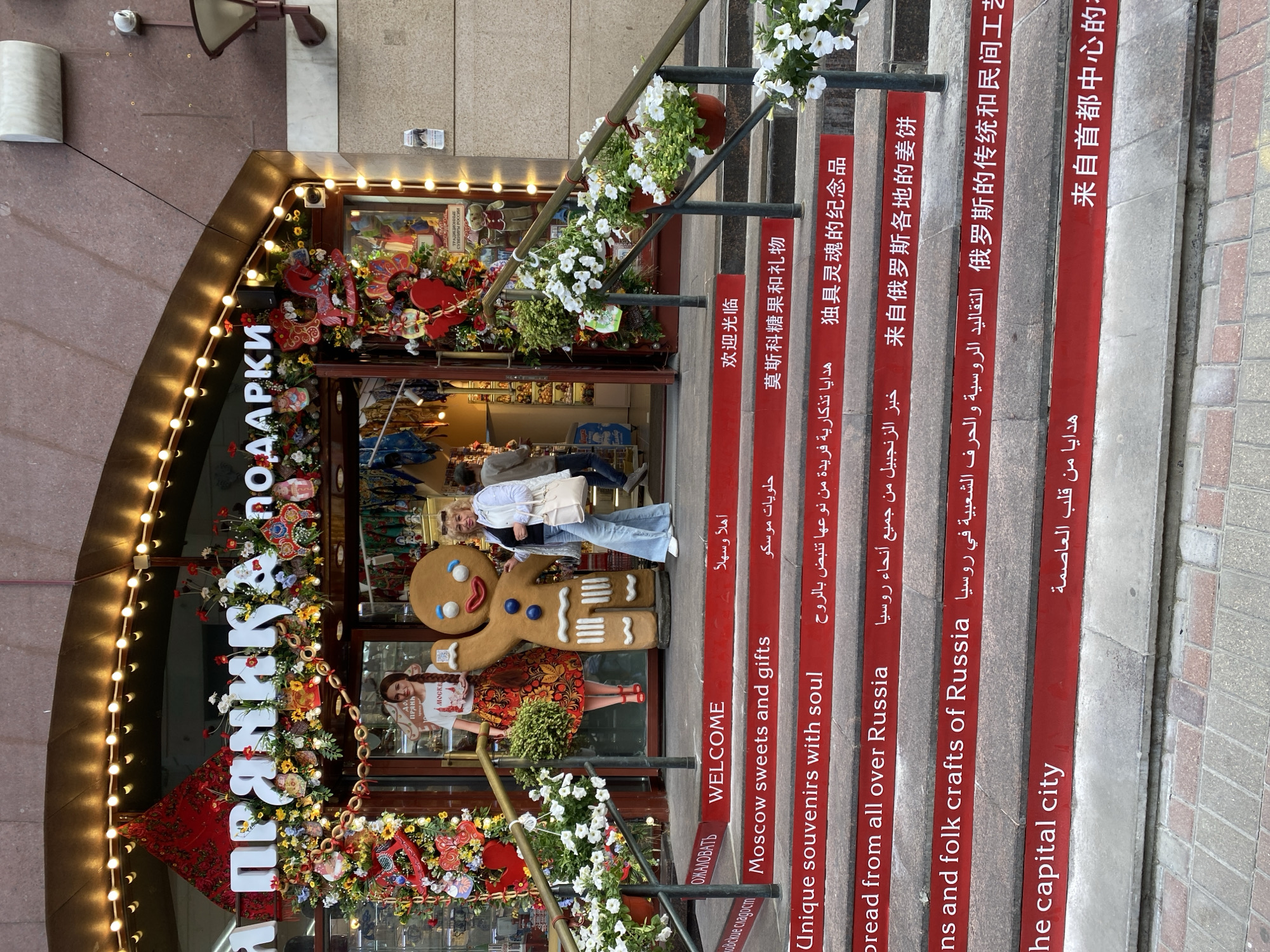 The image shows the entrance to a souvenir shop in Moscow, Russia. The shop's name is "Пряники", meaning "Gingerbread". The entrance is decorated with flowers and lights, and there is a large gingerbread man figure. A woman stands in front of the shop. The steps leading up to the entrance are painted with greetings in multiple languages, including English, Russian, Arabic, and Chinese. The translations relate to the store's theme, mentioning souvenirs, gingerbread, and Russian folk crafts.