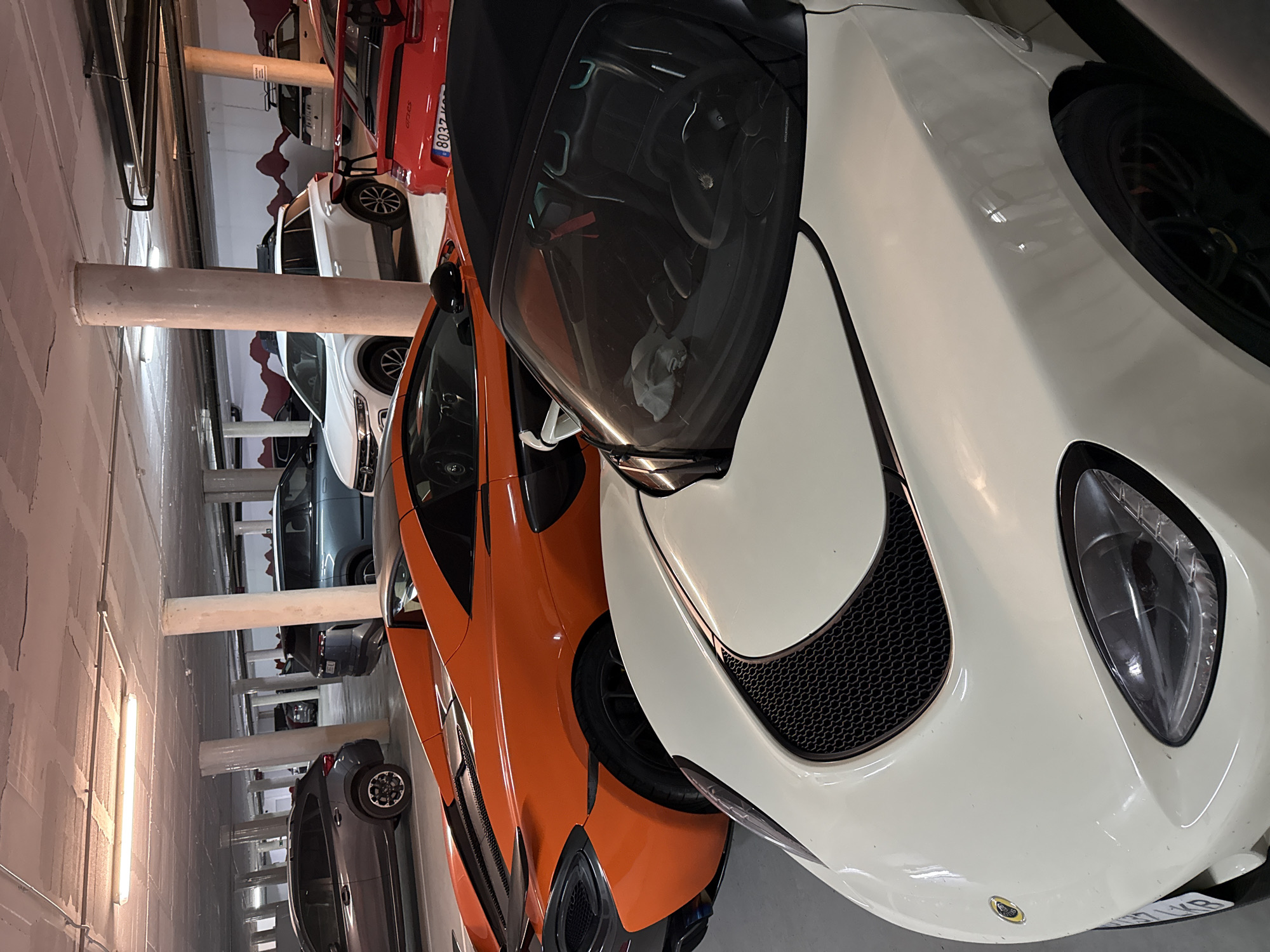 The image shows a close-up view of the front of two sports cars parked in a parking garage. The car on the right is white with a visible Lotus emblem. The car on the left is orange. The garage is brightly lit with fluorescent lights, and several other cars are visible in the background. The presence of a European license plate suggests this is Europe and the existence of the lotus emblems means the location is not in Benasque, Spain. The image lacks evidence of people or specific actions taking place.