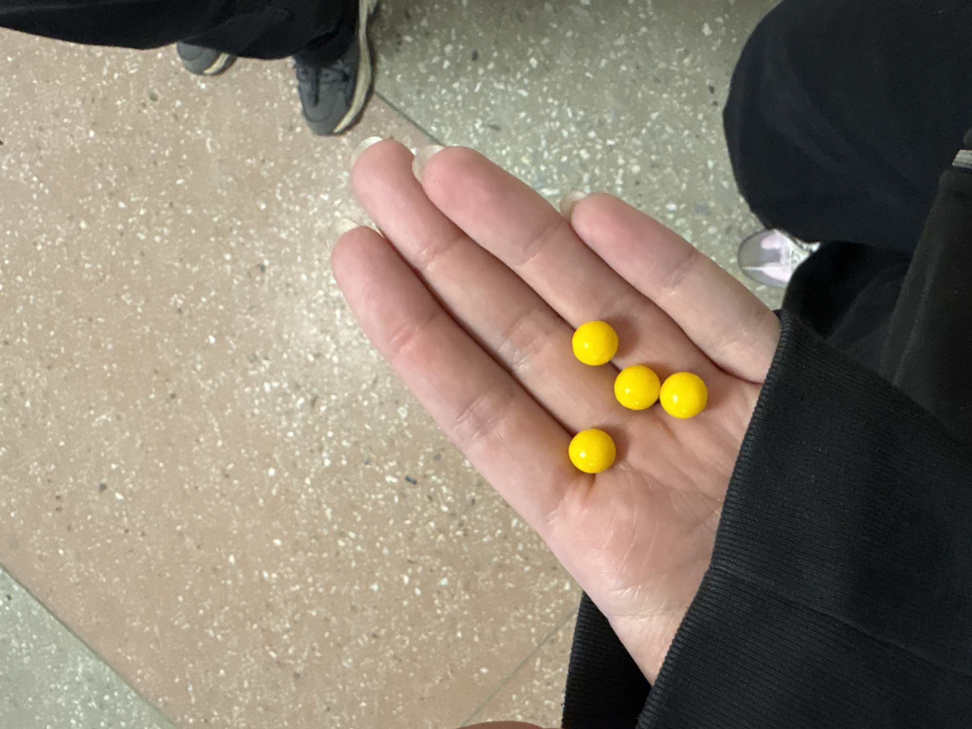 A pale-skinned human hand, likely female, holds four yellow spherical candies in its palm. The hand is positioned against a background that includes parts of the person's clothing (black jacket) and parts of the ground (beige speckled flooring). Other body parts are partially visible as well as other people who are not the main subject. There is not sufficient information to determine the city of Chaikovsky, Russia.