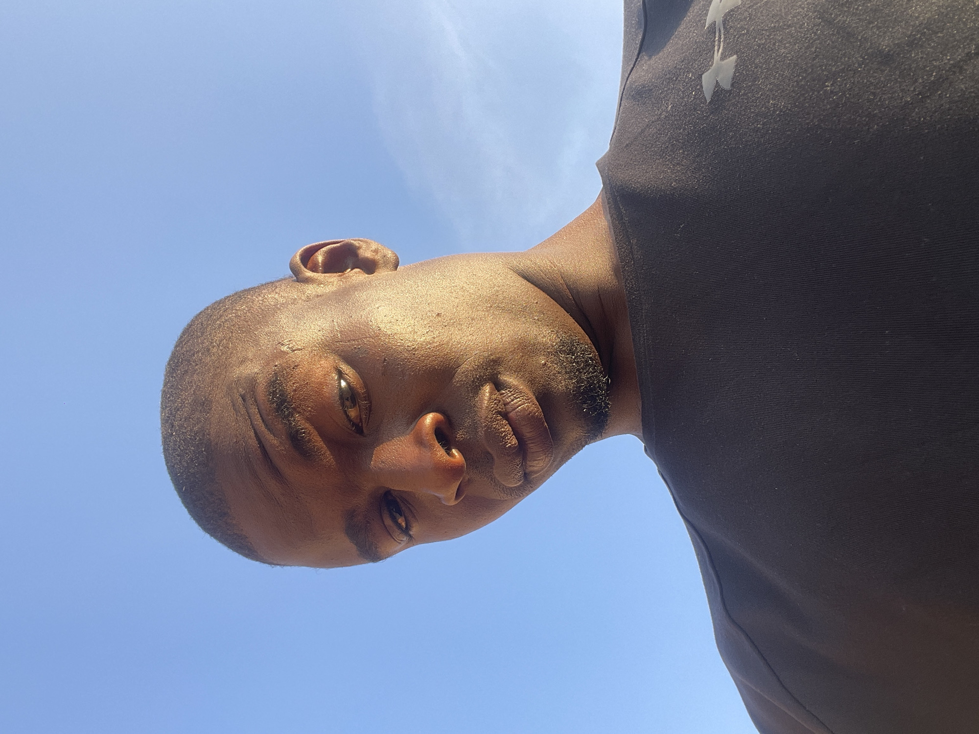 A low-angle headshot of a dark-skinned man against a blue sky with wispy clouds. The man has short hair and a close-cut beard. He's wearing a black Under Armour shirt. The photo appears to be taken outdoors in daylight. There is no indication of it being taken in Durimi, Nigeria.