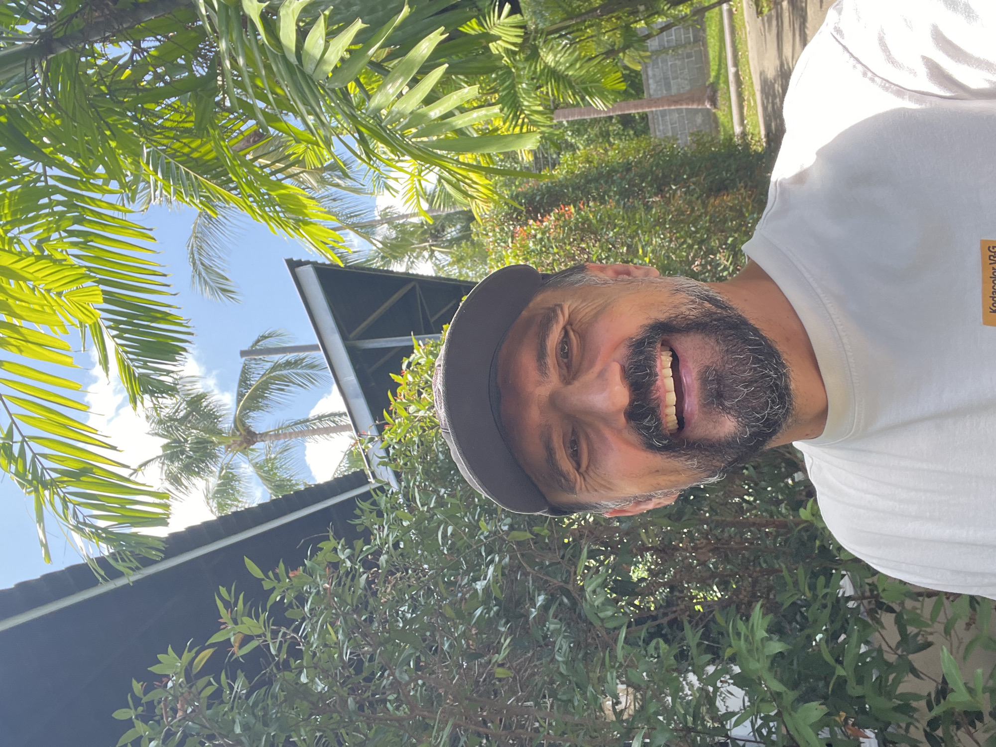The image is a low-angle selfie of a man with graying hair and a beard, wearing a cap and a white t-shirt, smiling broadly. The background shows lush green foliage including palm trees, and a building with a dark-colored roof can be seen in the midground. The location appears to be outdoors in a tropical climate, specifically บ้านบางรักษ์, Thailand, based on prior information.