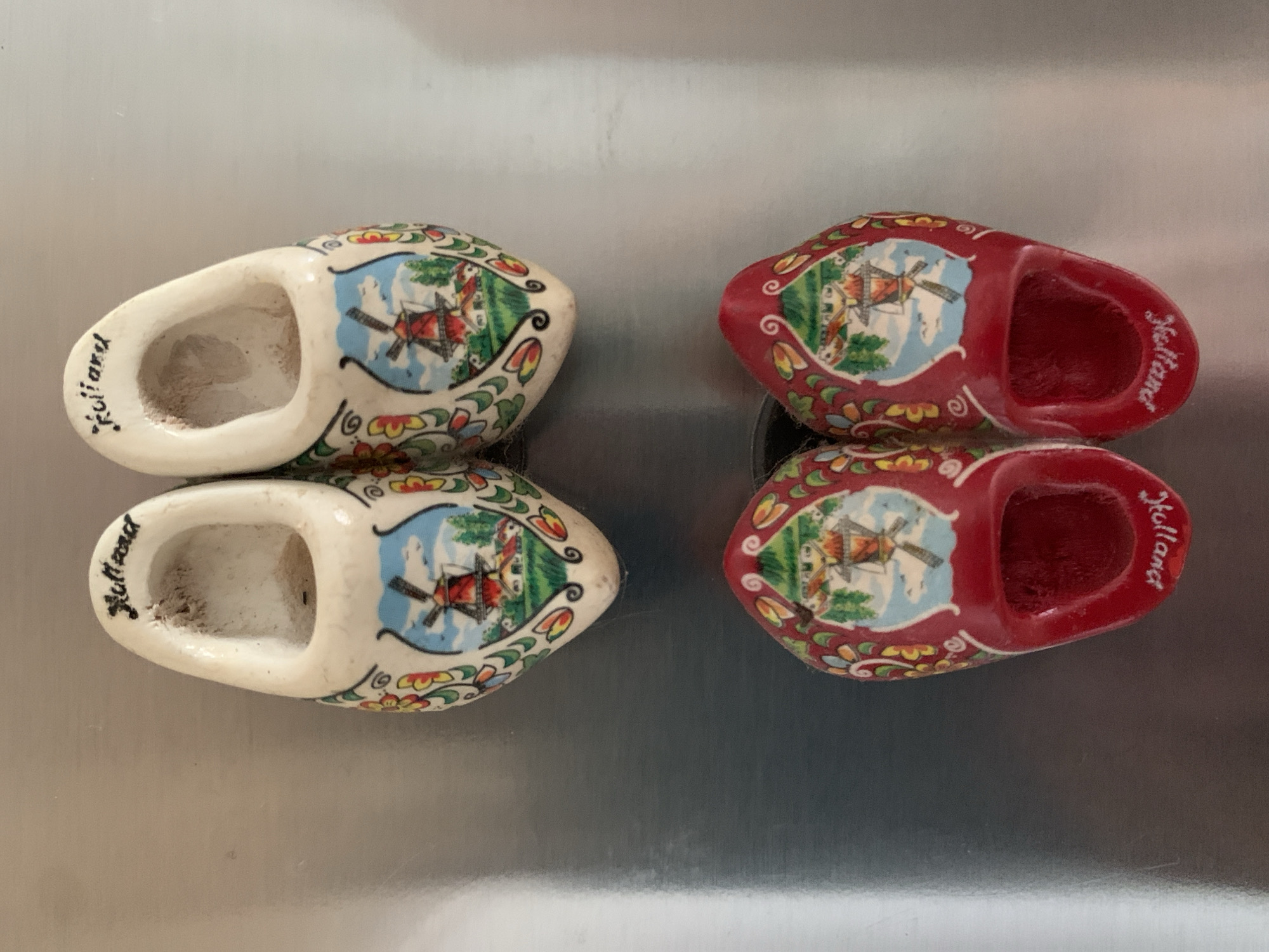 The image shows four miniature wooden clogs, which are traditional Dutch footwear. Two clogs are white and two are red, all adorned with painted scenes of windmills and floral patterns. The word "Holland" is written in script on the side of each clog. The clogs are arranged in two pairs, with the white clogs on the left and the red clogs on the right. They appear to be decorative items, possibly magnets or souvenirs, resting on a reflective, metallic surface. The lighting suggests the photo was taken indoors, with no specific time of day or weather conditions discernible. There are no people or any activity taking place in the image. The location context of Ivanava, Belarus, is external information and not visually evident from the image itself.