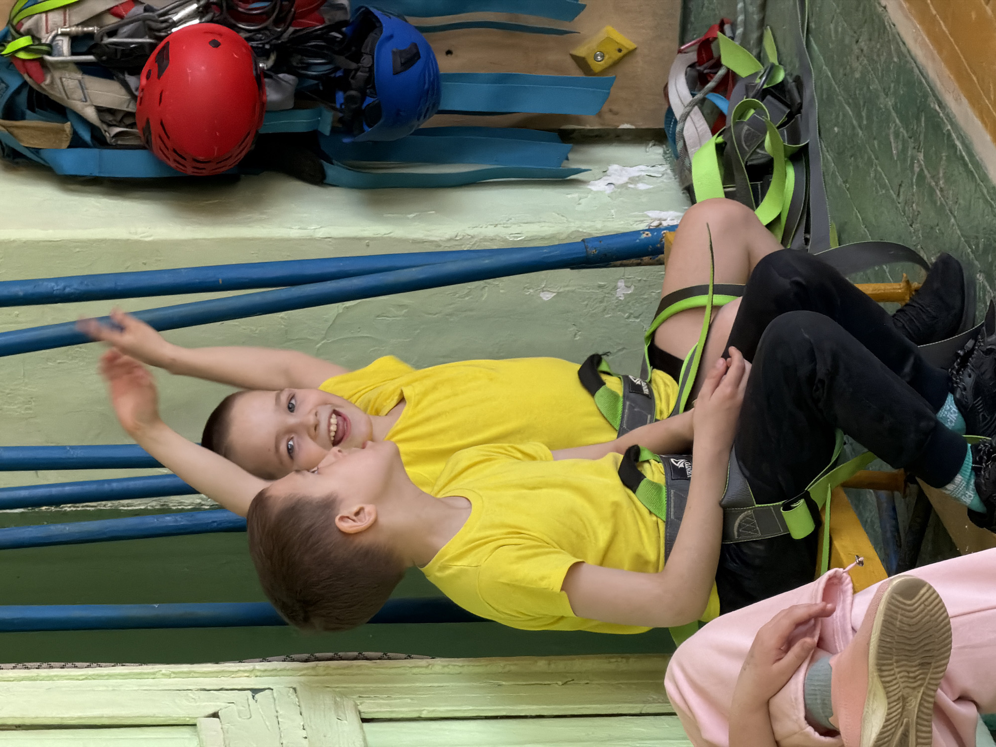 Two young boys, likely between 6 and 10 years old, are the primary subjects in this indoor image. Both are wearing identical bright yellow t-shirts and black pants, along with green and black climbing harnesses. The boy in the foreground has light brown hair and a soft smile, looking towards the boy behind him. The second boy, with fair skin and blue eyes, displays a wide, joyful smile, revealing his teeth, and is looking towards the camera. A third individual is partially visible in the bottom right, wearing a pink tracksuit, light blue socks, and a light-colored athletic shoe.
The setting appears to be a climbing gym or a sports facility equipped for climbing activities. In the background, various climbing gear is neatly stored on a shelf or hung on a wall: a red climbing helmet, a blue climbing helmet, ropes, carabiners, and additional harnesses. Blue metal bars run horizontally across the midground. The boys' harnesses are branded "CAMP," visible on the black straps.
The boys are lying on their backs, possibly resting or playing on the floor or a low platform after a climbing session. Their joyful expressions suggest a happy and relaxed atmosphere. The facility has light green and cream-colored painted walls, with some signs of wear. The image provides no specific time of day or weather cues, being an indoor scene in Izhevsk, Russia.
