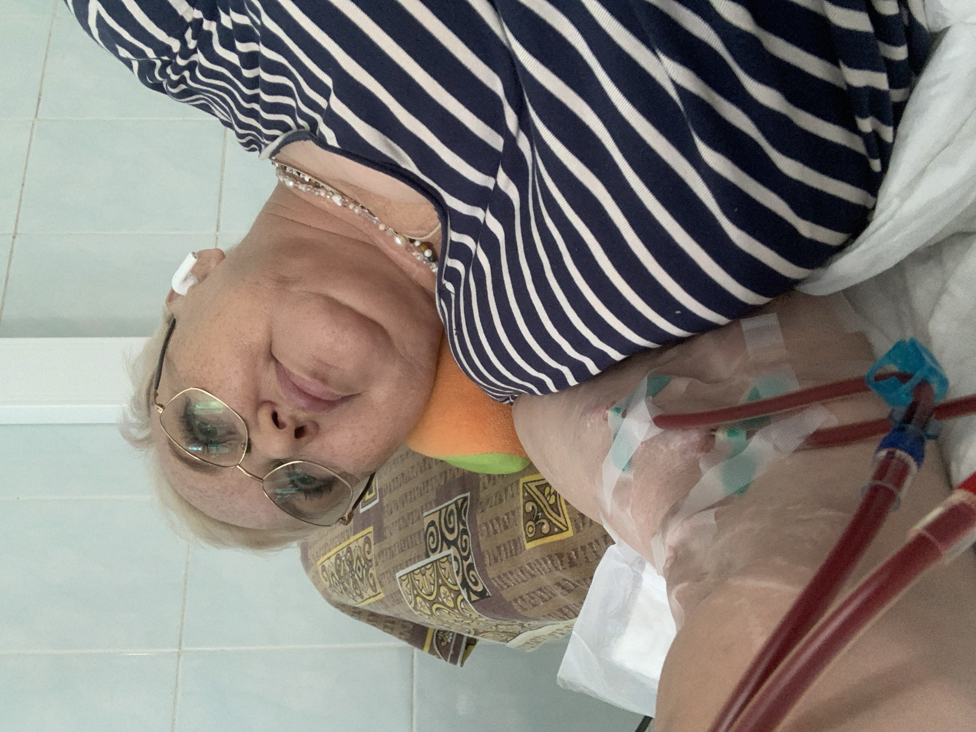 An adult female is lying on her side in a bed with a white sheet, wearing a striped blue and white shirt, a pearl necklace, and gold-rimmed glasses. A medical device with red tubes is taped to her chest. She has a white hearing aid in her left ear and is resting her head on an orange and green pillow. The background is a light blue tiled wall. The location in the city of Chaikovsky, Russia is not verifiable from the image.