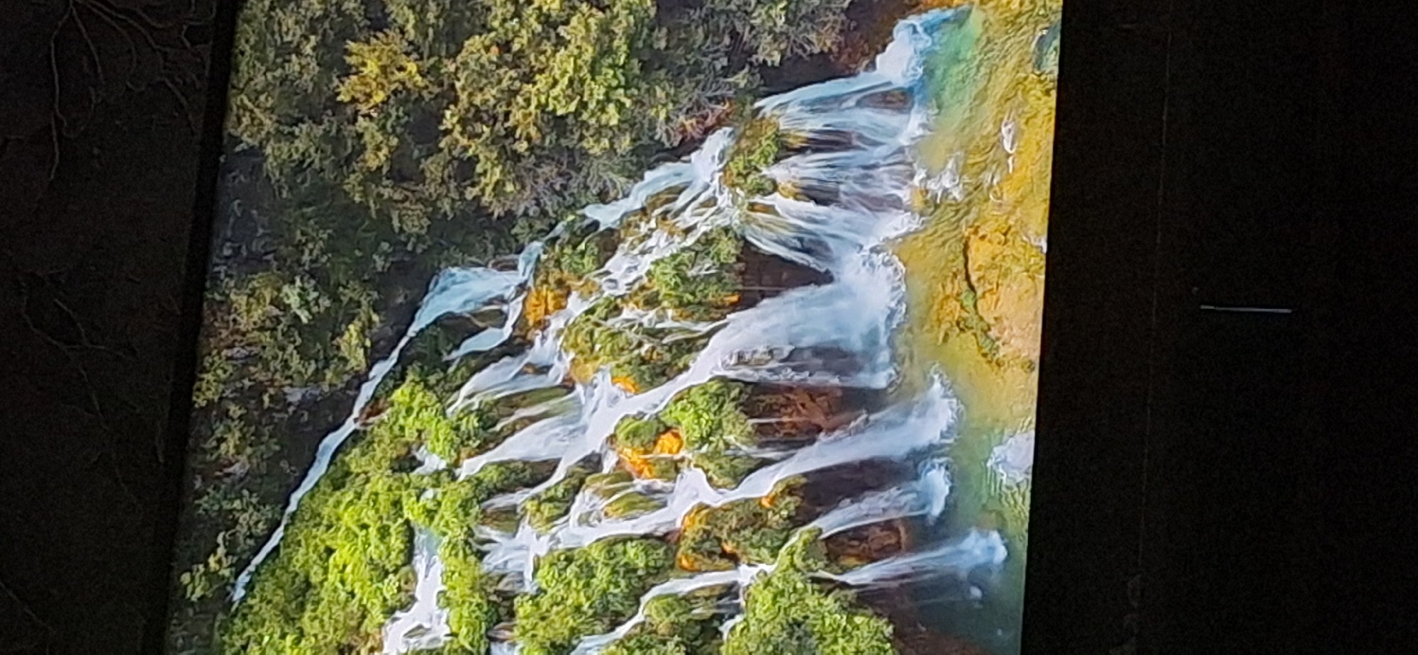 The image depicts a terraced waterfall cascading down a lush green hillside, dotted with rocky outcroppings and trees. The water is a milky white, creating a frothy texture as it flows over the rocks and into a shallow, yellowish-green pool at the base. The vegetation is dense and vibrant, with patches of bright green foliage contrasting with the brown and orange hues of the rocks. The top of the image shows the dark, textured surface of what appears to be a television screen or monitor, suggesting the image is a photograph of a display. The bottom of the image is predominantly black, likely representing the bezel or surrounding area of the display. There are no people, objects, or discernible actions other than the natural flow of the waterfall. The scene does not contain any identifiable landmarks or elements that would specifically place it within the city of Bila Tserkva, Ukraine.