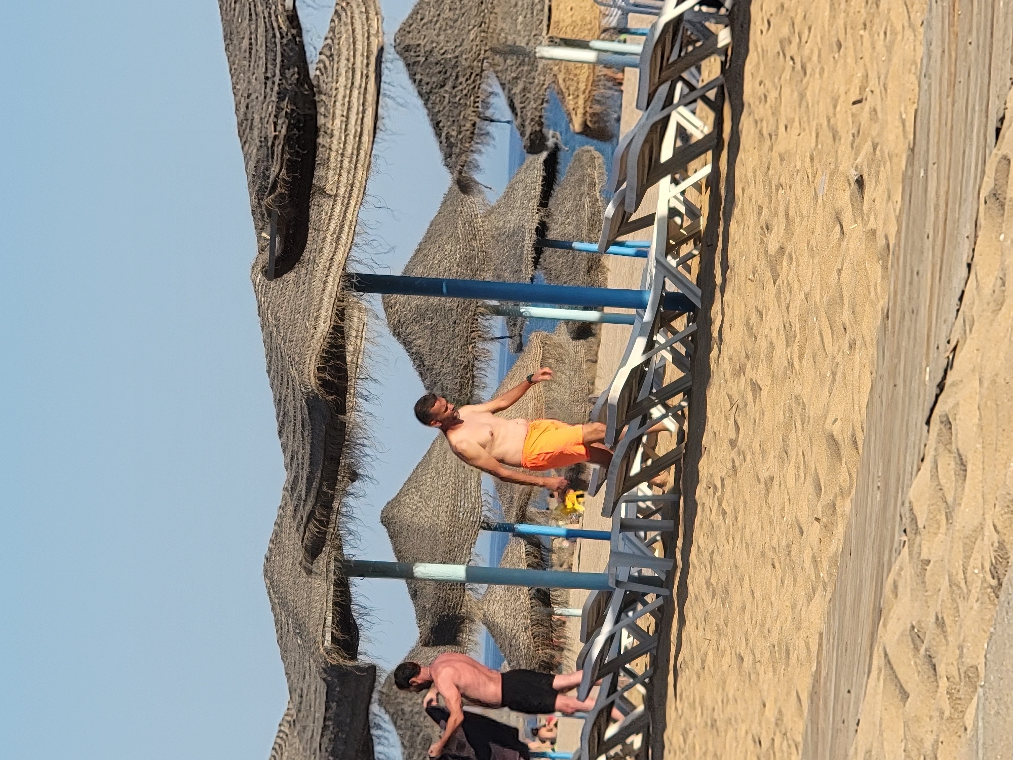 The image shows two men on a beach in Quarteira, Portugal. The scene is set with thatched sunshades and rows of lounge chairs, typical of a beach resort. The sand is golden and appears well-raked, with the ocean visible in the background under a clear blue sky.
The first man, in the foreground and closer to the camera, is wearing black swim shorts and is bent over, possibly adjusting something on the sand or a lounge chair. He has dark hair and a muscular build.
Further back and to the left, another man is seen standing and leaning on a lounge chair. He is wearing bright orange swim shorts and his torso is bare. He is looking off to the side and appears to be in a relaxed pose. A yellow object, possibly a cup or small bucket, is near his hand.
The sun is bright, casting distinct shadows, suggesting it is daytime, likely midday or early afternoon given the angle of the light. The overall mood of the image is one of leisure and relaxation on a sunny day at the beach. There is no visible text in the image.