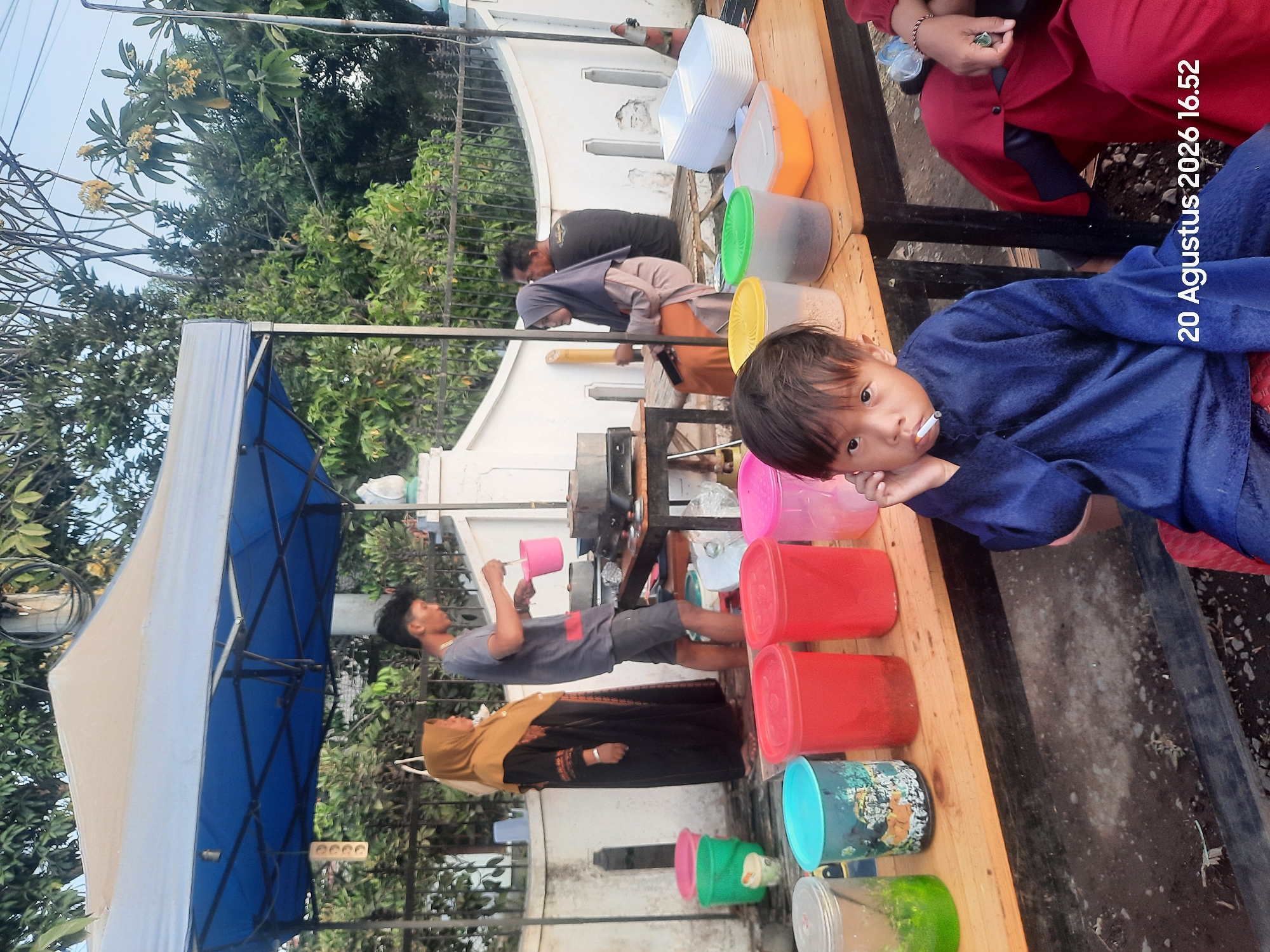 In the city of Sukokerto, Indonesia, a scene unfolds outdoors under a blue and tan tent. A young boy, dressed in a dark blue tunic, sits in the foreground, holding a cigarette to his lips. To his right, a person in a red garment with a patterned maroon hijab is partially visible, their right hand adorned with jewelry.
Behind them, a wooden table displays an array of plastic containers, some filled with colored liquids and others appearing empty. Among these are red, pink, and blue containers. Further back, a man in a gray t-shirt and dark shorts is actively preparing something, holding a pink bucket and a stirring utensil. Beside him, a woman in a brown hijab and a black and gold patterned dress stands observing. Another woman in a purple hijab and an orange skirt sits on a bench.
The background reveals a white wall with vertical decorative elements, a black metal fence, and lush green trees. The ground is a mix of dirt and gravel. A date and time stamp, "20 Augustus 2026 16.52," is visible in the lower right corner of the image.