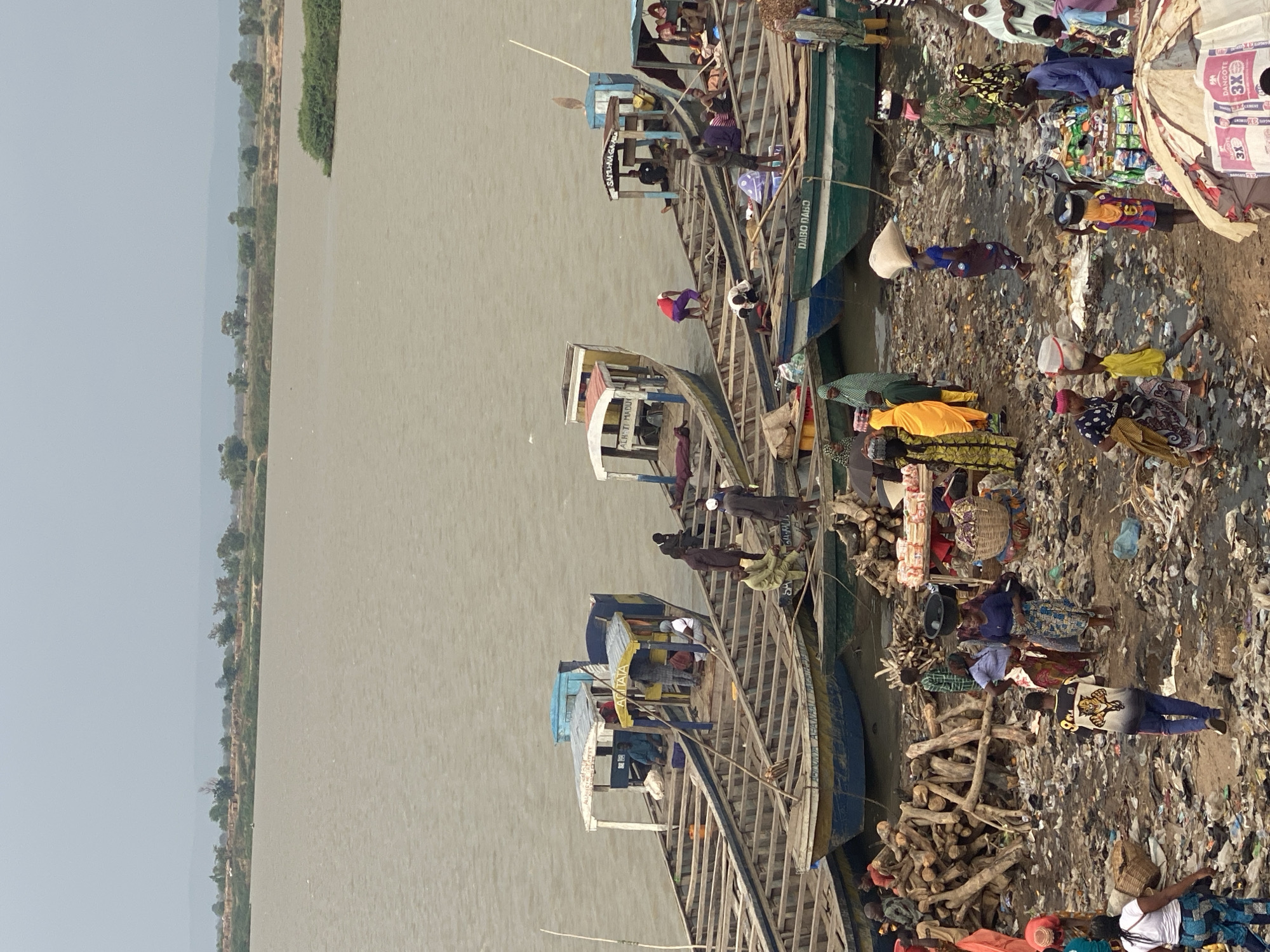 This image captures a bustling riverine scene, likely a market or transportation hub, set along a wide, murky river under a clear daytime sky. The background shows the expansive river stretching into the distance, bordered by a faint treeline and hazy hills.
The midground features several long, wooden boats, resembling traditional canoes or barges, docked closely together along the bank. These boats have simple wooden structures and some include small, enclosed cabins or shelters. People are visible on and around the boats, suggesting activity related to travel or commerce. Visible text on the boats includes "DABO DABO", "SAYTU-N-GA AHMAN", "ALT TI MADDU", and "ACATATA".
The foreground depicts the riverbank, which is a hive of activity. The ground is a mix of dirt, sand, and scattered debris and litter. Numerous people are present, dressed in diverse and colorful traditional African clothing. They are engaged in various activities: some carry large sacks or baskets on their heads, others stand near makeshift market stalls displaying packaged goods, and groups converse or move through the crowded area. Piles of chopped firewood are also stacked on the bank. A white sack prominently displays "DANGOTE 3X CEMENT", indicating a local Nigerian product. The scene conveys a dynamic environment centered around river transport and trade.