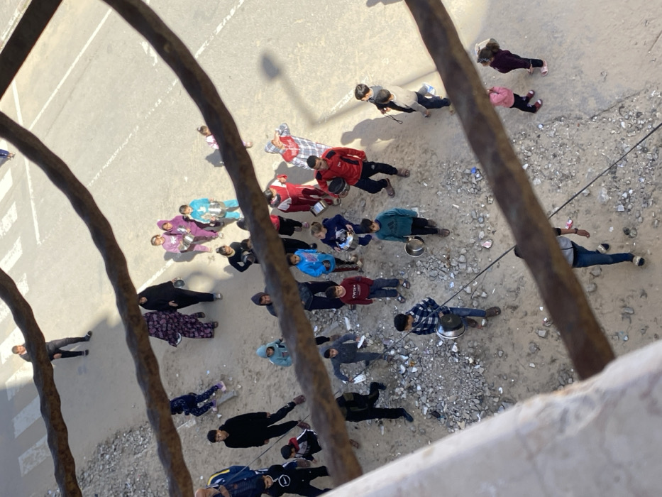 An overhead view from a structure in Khan Yunis, Palestinian Territory, frames a scene below through rusty metal bars. The ground surface transitions from a paved road with a visible white pedestrian crossing in the upper background and right, to an unpaved, debris-strewn dirt area in the center and foreground. Approximately 30 to 40 individuals, encompassing adults and numerous children of varying ages, are present. A significant number of these individuals, predominantly children, are carrying metal pots or bowls. People are observed walking and standing across both the paved and unpaved sections of the ground. Strong shadows are cast, indicating bright daylight conditions.