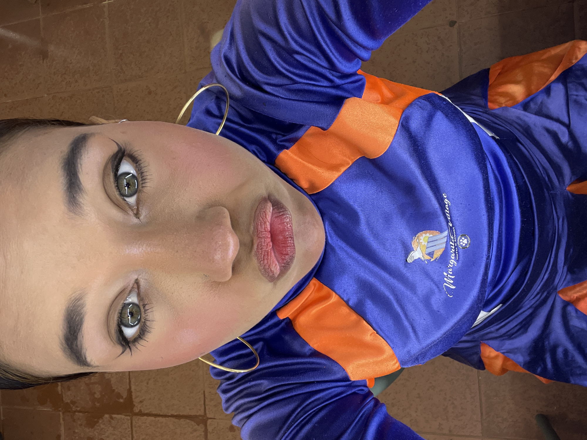 A close-up, low-angle shot depicts a person wearing a blue and orange satin-like jacket or jersey, presumably indoors against a tile floor. The person has fair skin, defined eyebrows, visible eyelashes, and large gold hoop earrings. The jacket is branded with the word "Margarita" and has an emblem associated with a resort on Margarita Island. The image suggests the person is located in Porlamar, Venezuela, due to the association of the Margarita Island resort with that region.