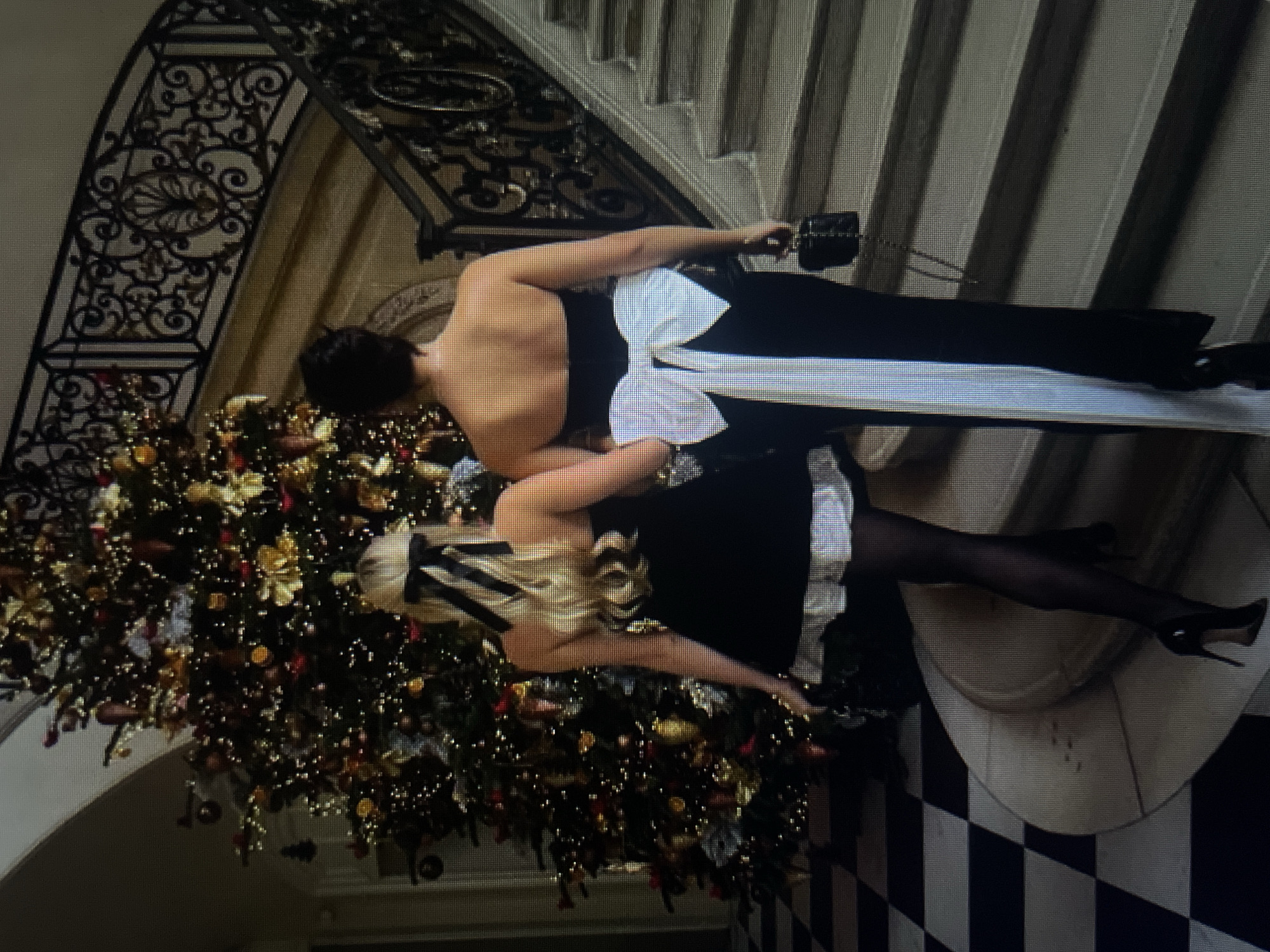 Two women are walking up a grand staircase, illuminated by a large, decorated Christmas tree. The woman on the left has blonde hair tied with a black bow and wears a black strapless dress with a white trim. She is wearing black stockings and high heels. The woman on the right has dark hair and wears a black strapless gown with a large white bow at the back. She is carrying a small black purse. The staircase has an ornate black metal railing. The floor in the foreground is a checkered pattern of black and white tiles. The scene appears to be indoors, possibly in a luxurious residence or hotel lobby, decorated for the holidays. There is no information provided that suggests the location is in the city of Kołobrzeg, Poland.
