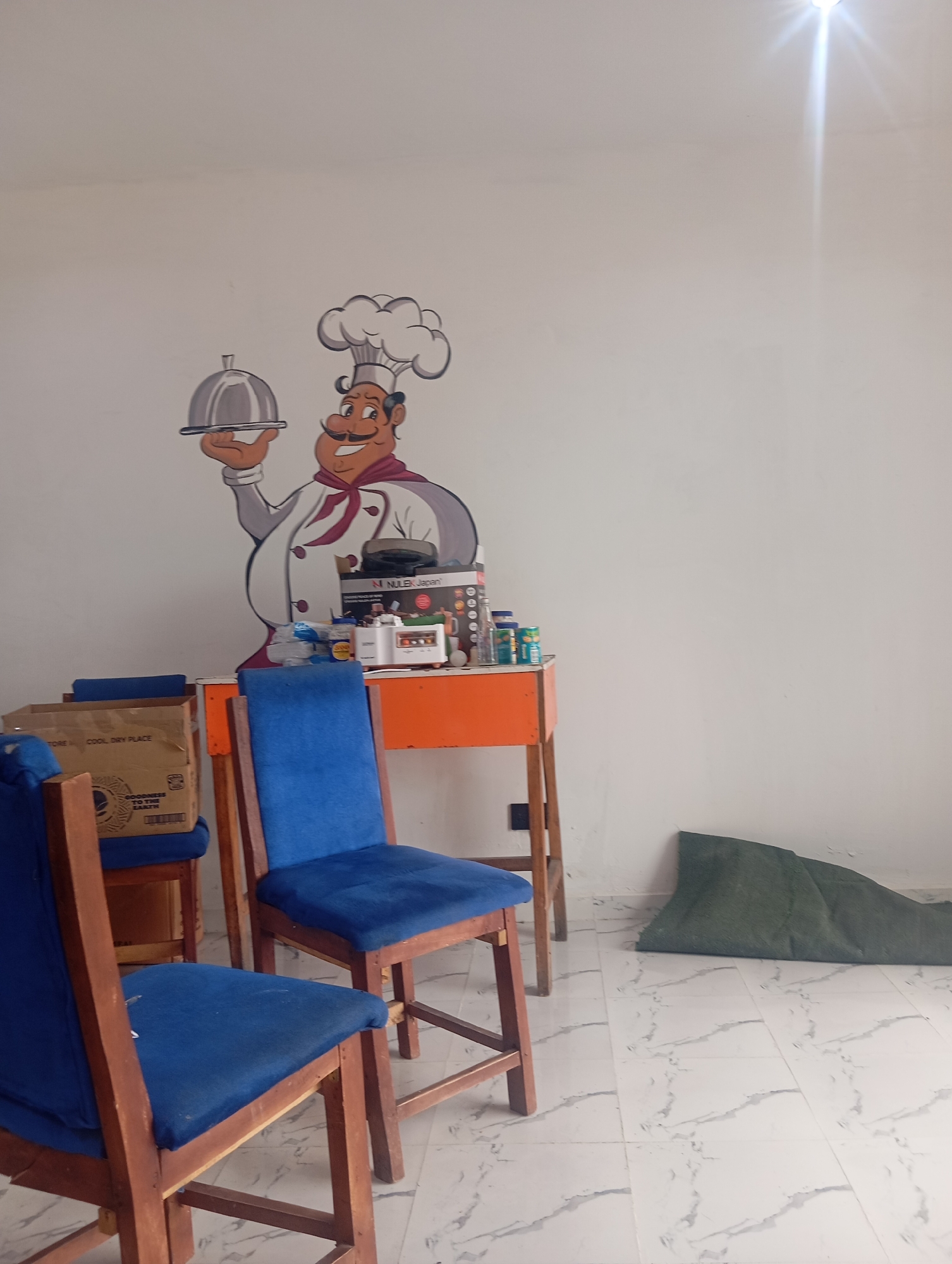 The image shows an interior scene in what appears to be a small restaurant or eatery. On the wall, there's a cartoon drawing of a chef holding a covered dish. An orange table is positioned beneath the drawing, cluttered with boxes of supplies and other items. Two blue cushioned chairs are positioned in front of the table. In the background, a rolled up green material is leaning against the wall. The floor is tiled with a white marble-like pattern. The location is believed to be in Gombe, Nigeria, based on prior image metadata.