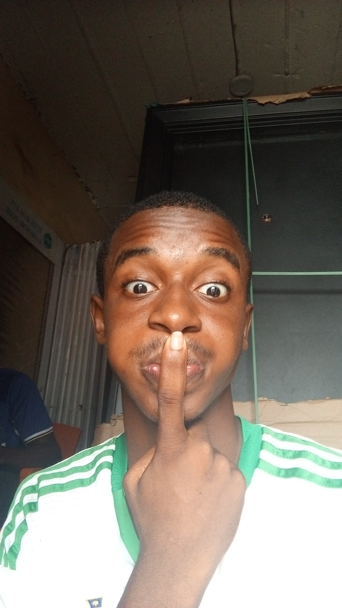 A selfie of a person with brown skin, a mustache, and short black hair is centered in the frame, positioned indoors with an upward angle. The individual is wearing a white shirt with green stripes on the sleeves and is holding their index finger vertically in front of their mouth, as if indicating silence. The expression on their face includes wide eyes and puffed cheeks.
Behind the person, there is a partially visible doorway on the right side of the frame, a corrugated surface on the left, and what appears to be a light fixture attached to the ceiling. The setting appears to be an indoor space with limited lighting. The location is Unknown, Unknown.