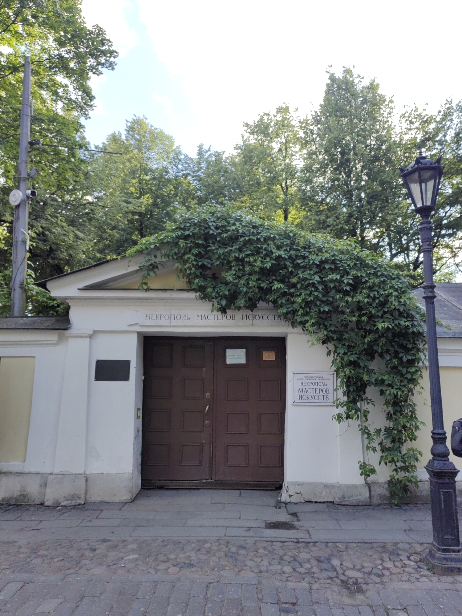The image shows the entrance to a necropolis, or cemetery, dedicated to masters of arts in Saint Petersburg, Russia. The building has a white facade with a prominent entrance featuring two large, dark brown double doors. Above the doors, a sign reads "НЕКРОПОЛЬ МАСТЕРОВ ИСКУССТВ" (Necropolis of Masters of Arts).
The entrance is partially covered by lush green ivy that cascades over the roof and down the side of the building. To the left of the doors, a small black plaque is affixed to the wall. To the right of the doors, another sign is mounted, also displaying the name "НЕКРОПОЛЬ МАСТЕРОВ ИСКУССТВ" and some smaller text above it which appears to be "ГОСУДАРСТВЕННЫЙ МУЗЕЙ ГОРОДСКОЙ СКУЛЬПТУРЫ" (State Museum of Urban Sculpture).
A traditional black lamppost stands to the right of the entrance, adding to the historic ambiance of the location. The ground in front of the building is paved with cobblestones. Trees with green foliage are visible behind and around the building, suggesting a park-like setting. A utility pole with some equipment attached is visible on the far left. The sky is bright, indicating it is daytime, likely a sunny or partly cloudy day. There are no people visible in the image. The overall impression is one of a well-maintained historical site.