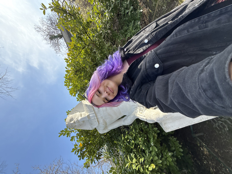 The image displays a young adult female from the torso up, taking a selfie outdoors. She possesses long, wavy hair that transitions from pink at the front to vibrant purple, and she exhibits a faint smile while looking directly into the camera. She is dressed in an oversized black denim jacket featuring a dark faux-shearling collar, worn over a maroon top, with sunglasses perched on her head. To her left and slightly behind her stands a white stone statue depicting an ancient Egyptian pharaoh, characterized by a nemes headdress, false beard, and a kilt. Both the individual and the statue are situated amidst lush green foliage from shrubs and trees. The background features a clear blue sky with sparse white clouds, bare tree branches, and a partial building structure. The scene is illuminated by natural sunlight, casting soft shadows. This location is identified as the Botanical Garden of the Faculty of Science, University of Zagreb, in the City of Zagreb, Croatia.