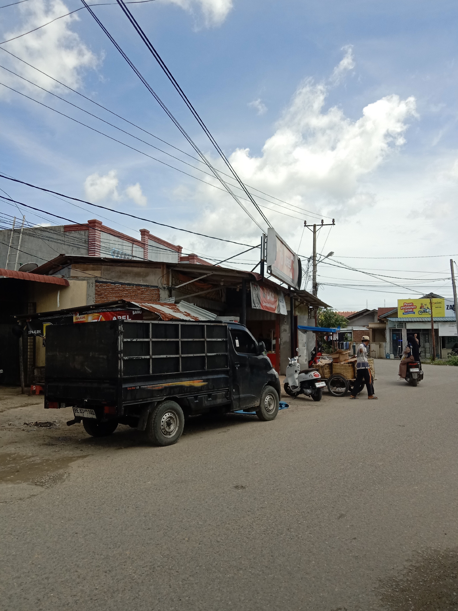 The image shows a street scene in Lhokseumawe, Indonesia. On the left, a black pick-up truck is parked, partially blocking the road. Nearby, a person pulls a cart with items on it. Further down the road, there are two scooters and another person on a scooter. Buildings line the street, with signs visible on some of them. Power lines crisscross overhead against a blue sky with scattered clouds.