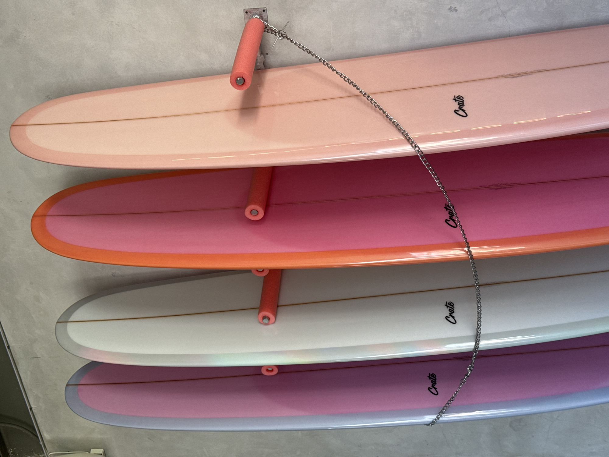 The image shows a close-up view of four surfboards stacked horizontally in a rack. The surfboards are different colors, including peach, pink and orange, white and purple. Each board has the word "Crate" written on it in black lettering. The boards are held in place by a rack system with orange foam padding and a metal chain attached to the ceiling. The background appears to be a gray concrete wall. The location in the city of Mengwi, Indonesia is not confirmed based on visual data.