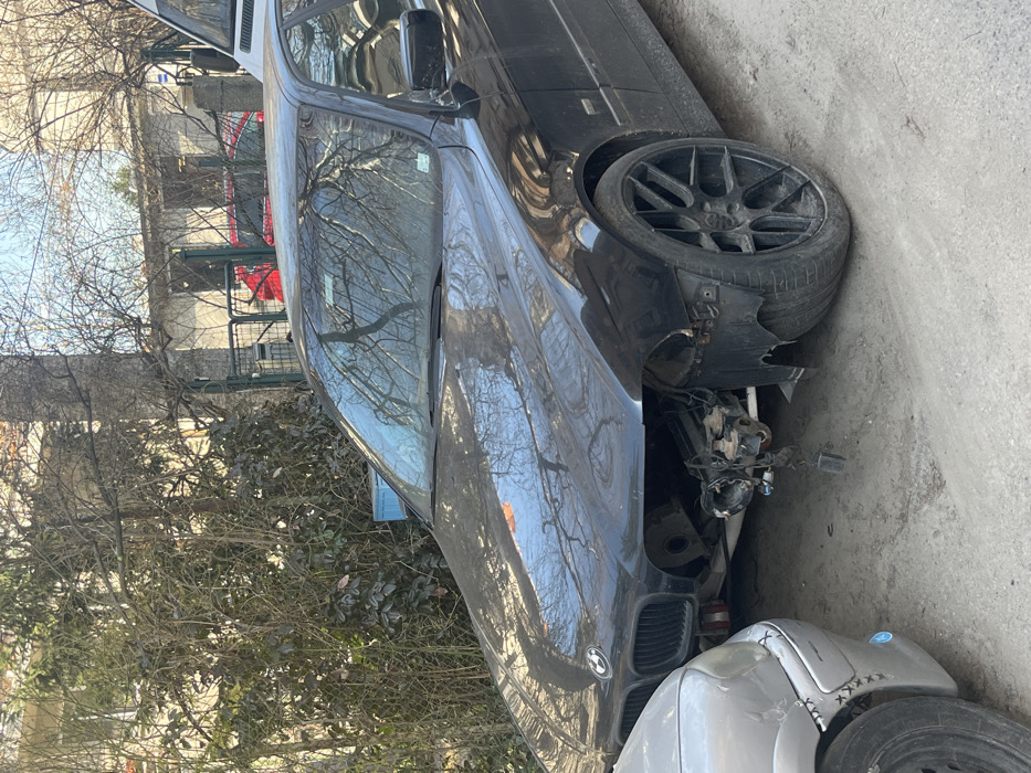 The image depicts an outdoor scene in what appears to be a residential area of Sofia, Bulgaria, characterized by an unpaved dirt and gravel street. Two vehicles are prominently featured. A black BMW E39 5-series sedan occupies the central and right foreground, facing left. This vehicle exhibits significant frontal damage, with the entire front bumper, grille, both headlights, and the right front fender missing, exposing the engine compartment and various chassis components. The hood is present but sits above the exposed engine bay, reflecting tree branches. The left front wheel is fitted with a black multi-spoke aftermarket alloy rim and a low-profile tire. Positioned directly in front of the BMW, partially visible on the left, is the front section of a silver-grey vehicle. Its front bumper displays visible stitch-like repairs or ties on the left side. The background features dense, mostly bare deciduous trees and evergreen shrubs, partially obscuring light-colored, possibly stuccoed, residential buildings and a metal gate. The upper portion of a white van is visible on the far right, behind the BMW. A blue object, possibly a crate, is partially visible within the foliage on the left. No individuals are present in the scene.
