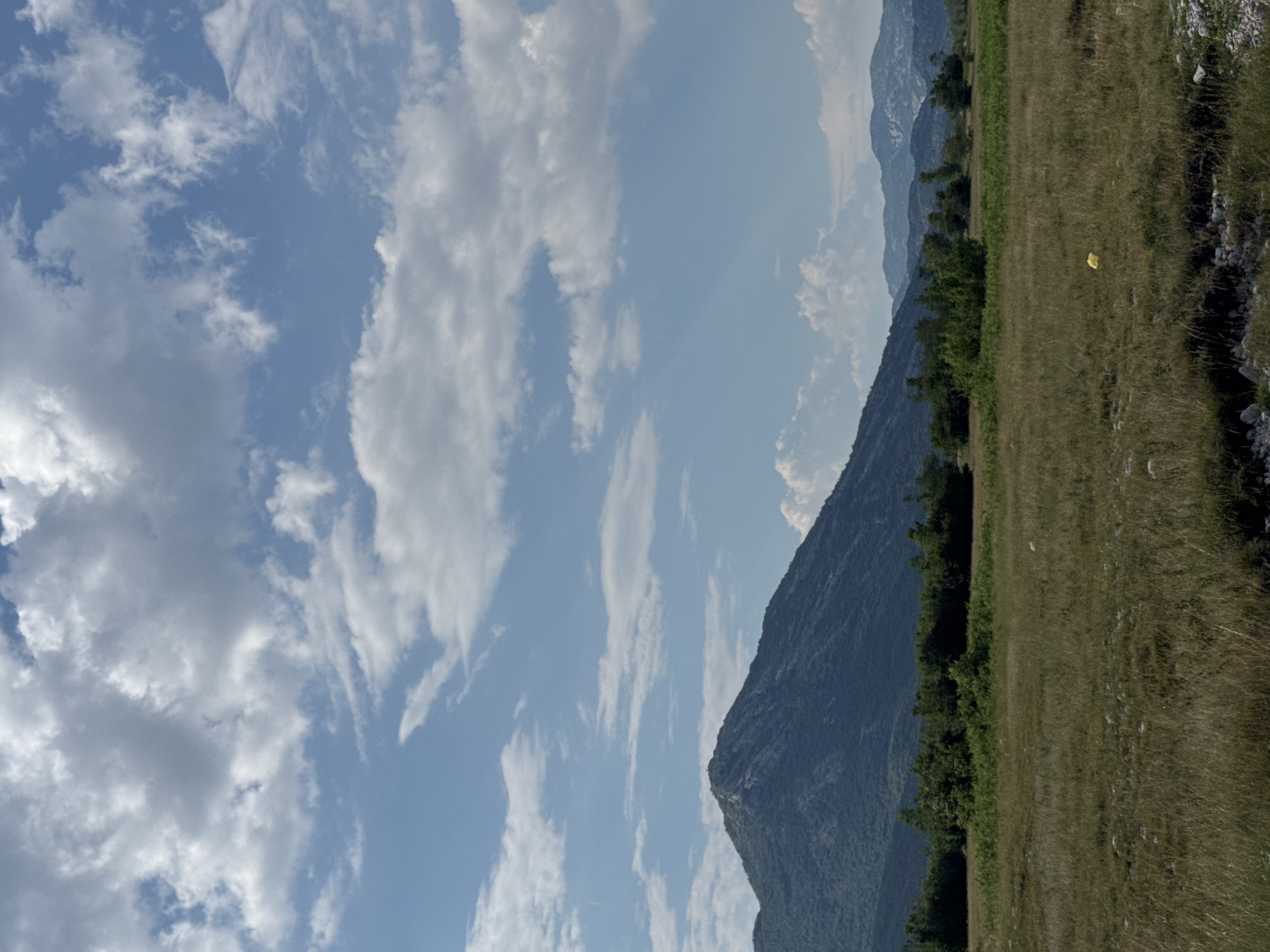 The image depicts a landscape in the vicinity of Dragalj, Montenegro. A prominent, tree-covered mountain dominates the middle ground, its slopes receding into the distance. In the foreground, a dry, grassy field stretches out, dotted with scattered rocks. A single yellow object, possibly a piece of debris or litter, is visible in the field. The sky above is a mixture of blue and white clouds, with some rays of sunlight breaking through. The overall scene conveys a sense of natural, undeveloped terrain.